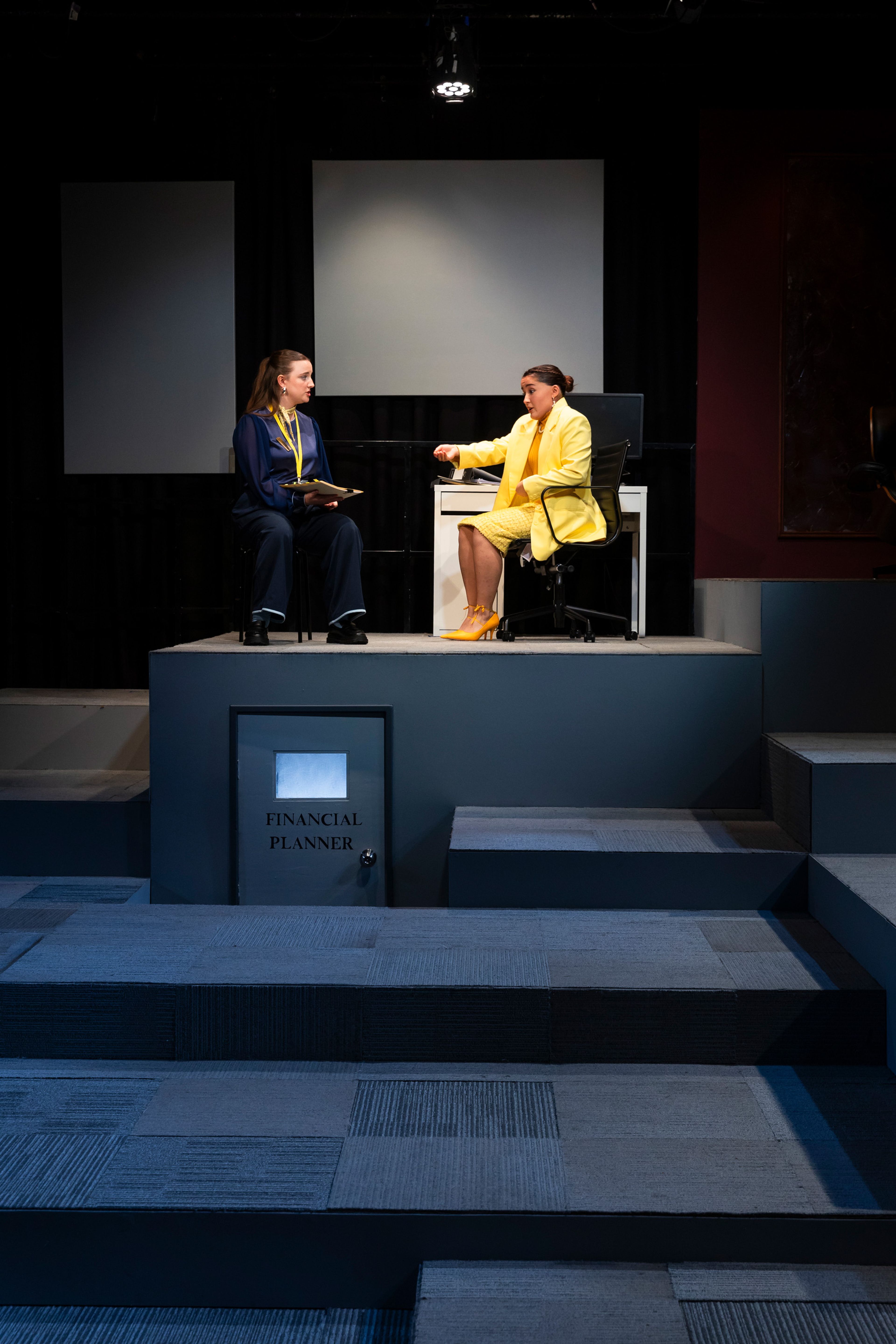 Theatre production photograph showing a scene in a financial planner's office, with two performers on a raised platform, one in navy blue and one in bright yellow, with a door labeled 'Financial Planner' visible below.