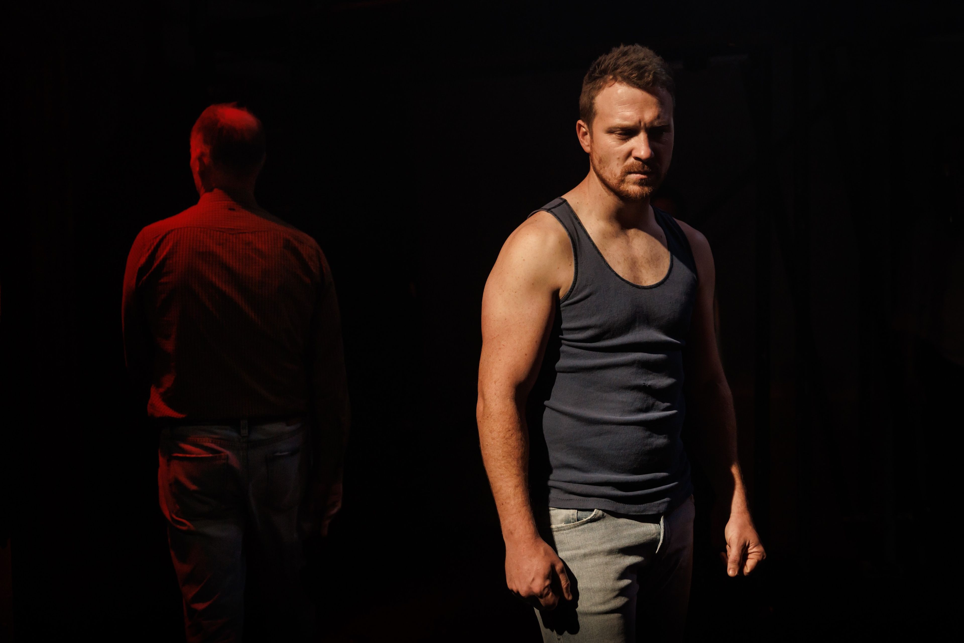 Theatre production photograph of a man in a dark tank top standing under dramatic lighting, with another figure partially visible in red light in the background.