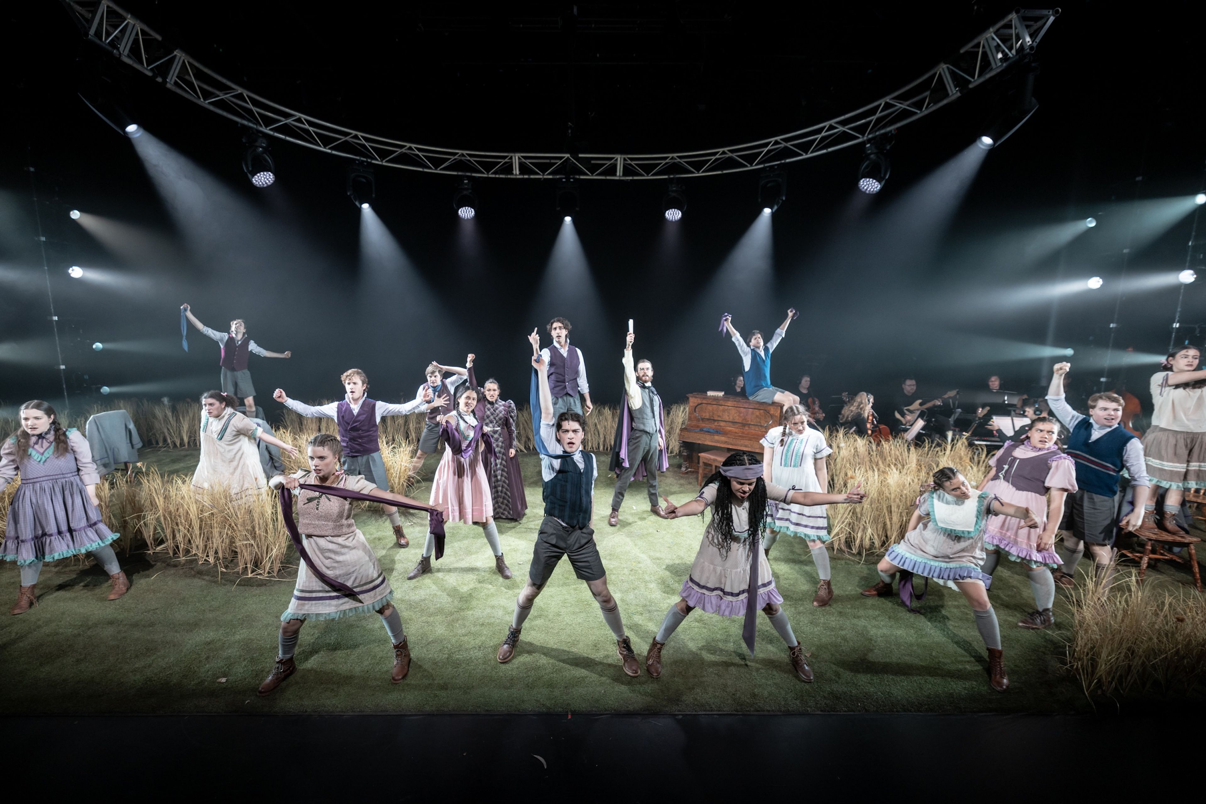 Theatre production photograph showing ensemble cast in period costumes performing energetic choreography on a stage set with tall grass, dramatic overhead lighting, and curved lighting rig visible.