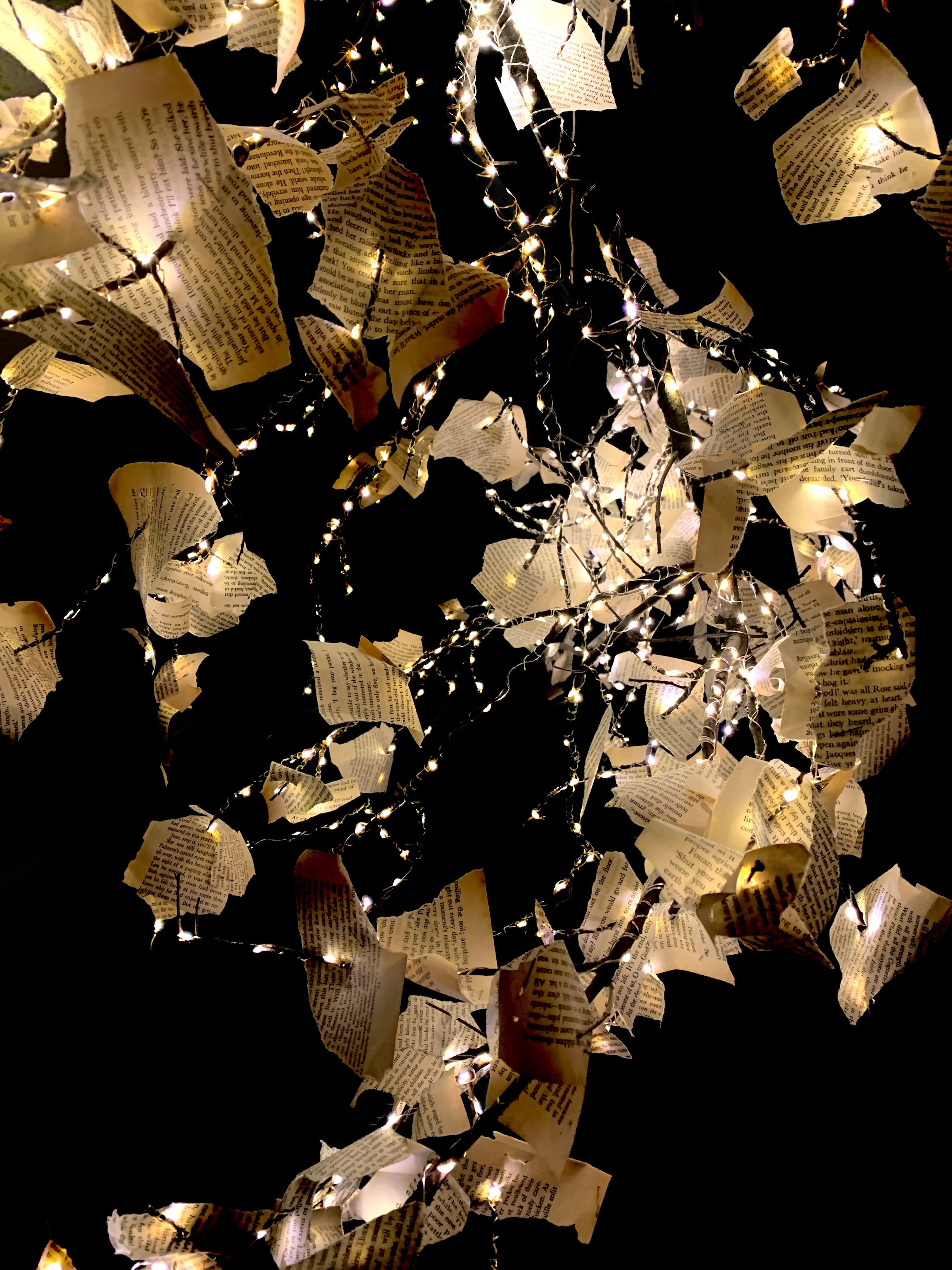 Close-up view of an installation featuring torn pages from a book intertwined with delicate string lights. The lights illuminate the pages, casting a warm, enchanting glow against a black background, highlighting the texture and text on each piece of paper.