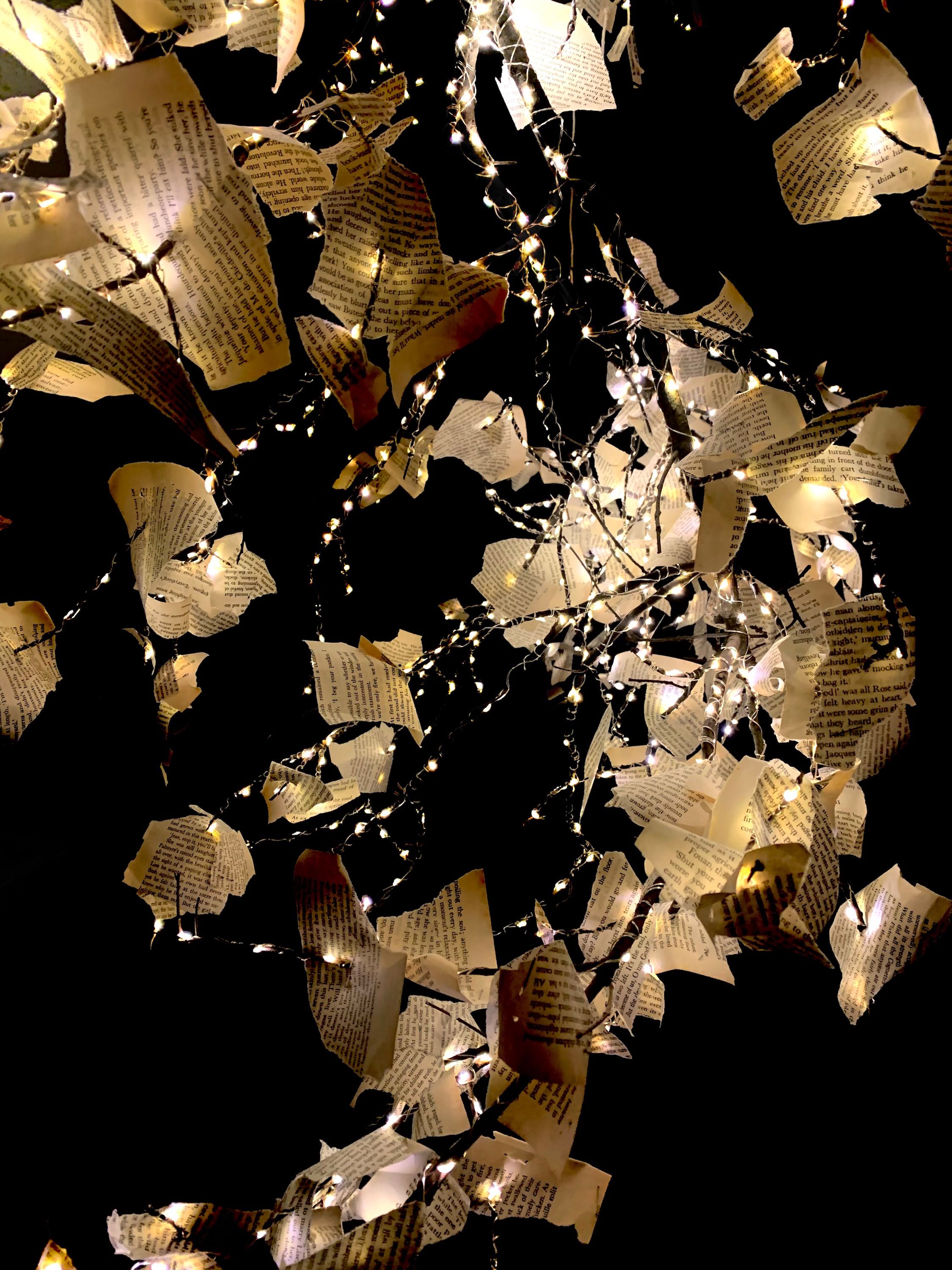 Close-up view of an installation featuring torn pages from a book intertwined with delicate string lights. The lights illuminate the pages, casting a warm, enchanting glow against a black background, highlighting the texture and text on each piece of paper.