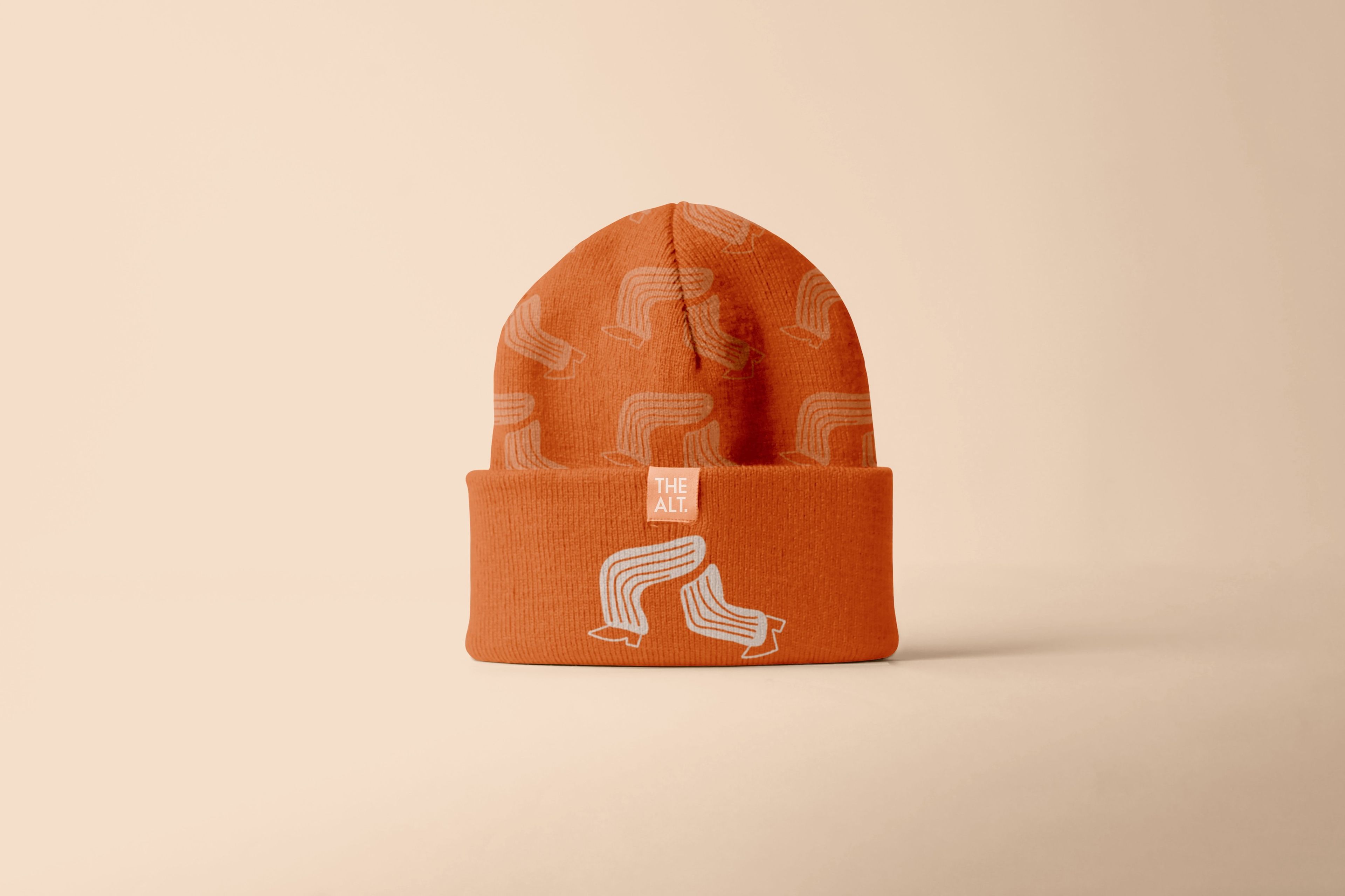 A close-up of an orange beanie labelled "THE ALT." The beanie has a repeating minimalist graphic of curved lines resembling a stylised, flowing letter "A" or wave, printed in a subtle tone-on-tone design.