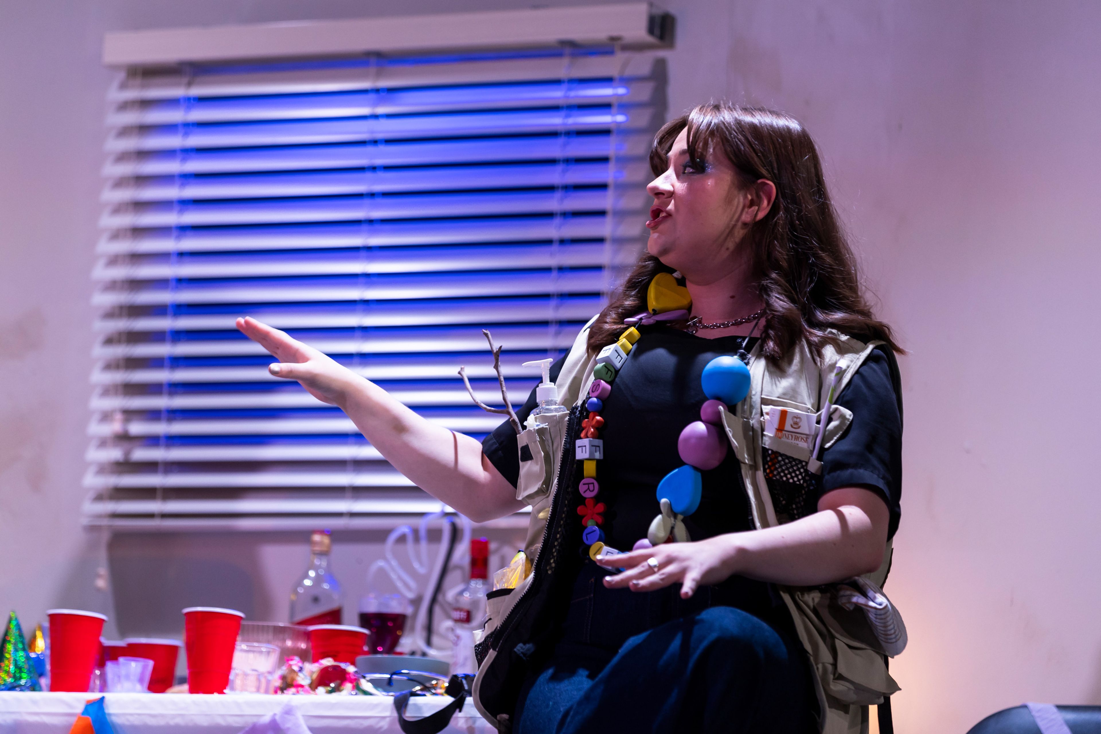 Theatre production photograph of a performer gesturing while speaking, wearing an oversized beaded necklace and vest, with blue-lit window blinds in the background.