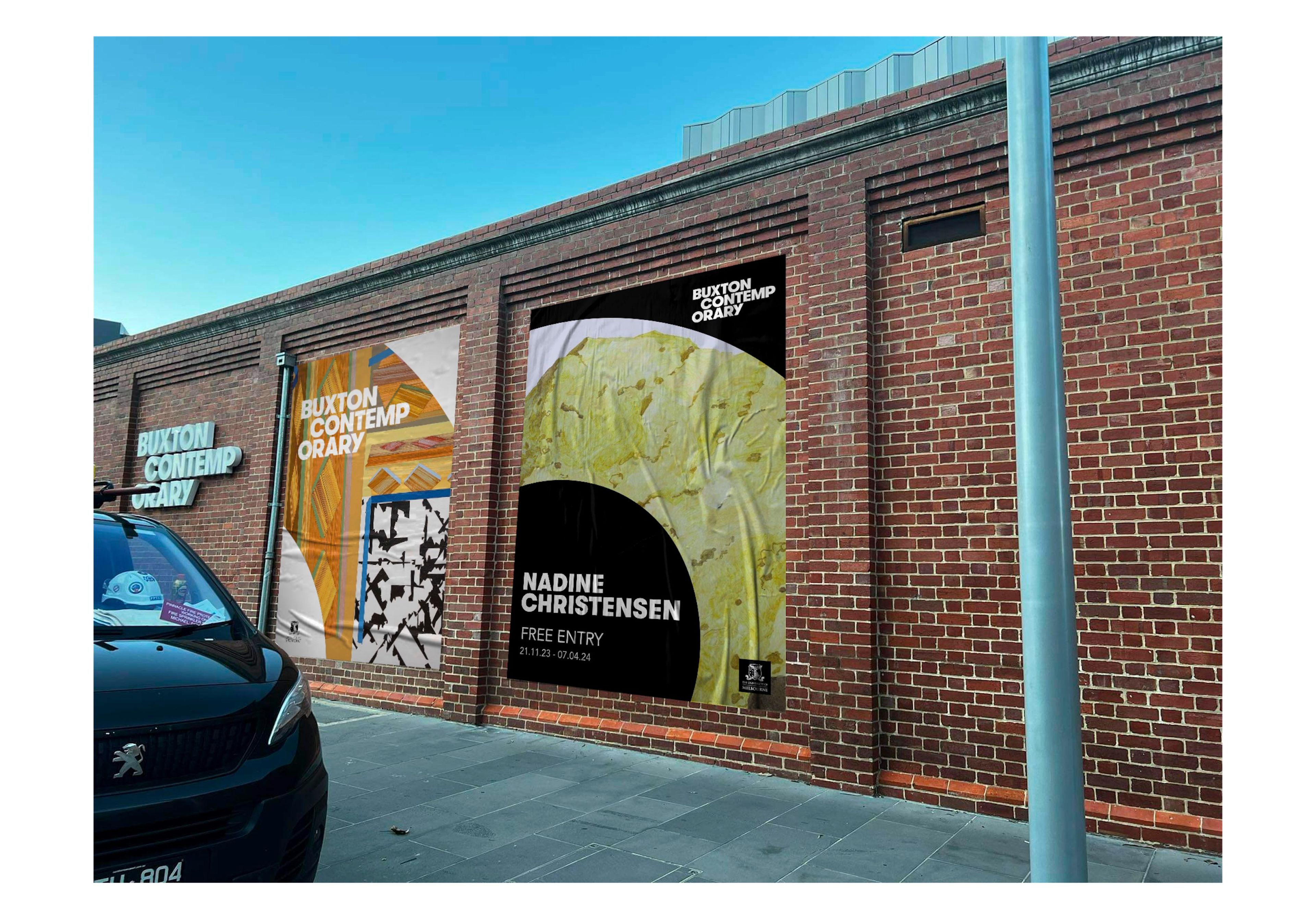 Exterior photograph of Buxton Contemporary's brick facade displaying three large-format exhibition banners. Featured poster for Nadine Christensen exhibition uses striking black and yellow design with exhibition dates.