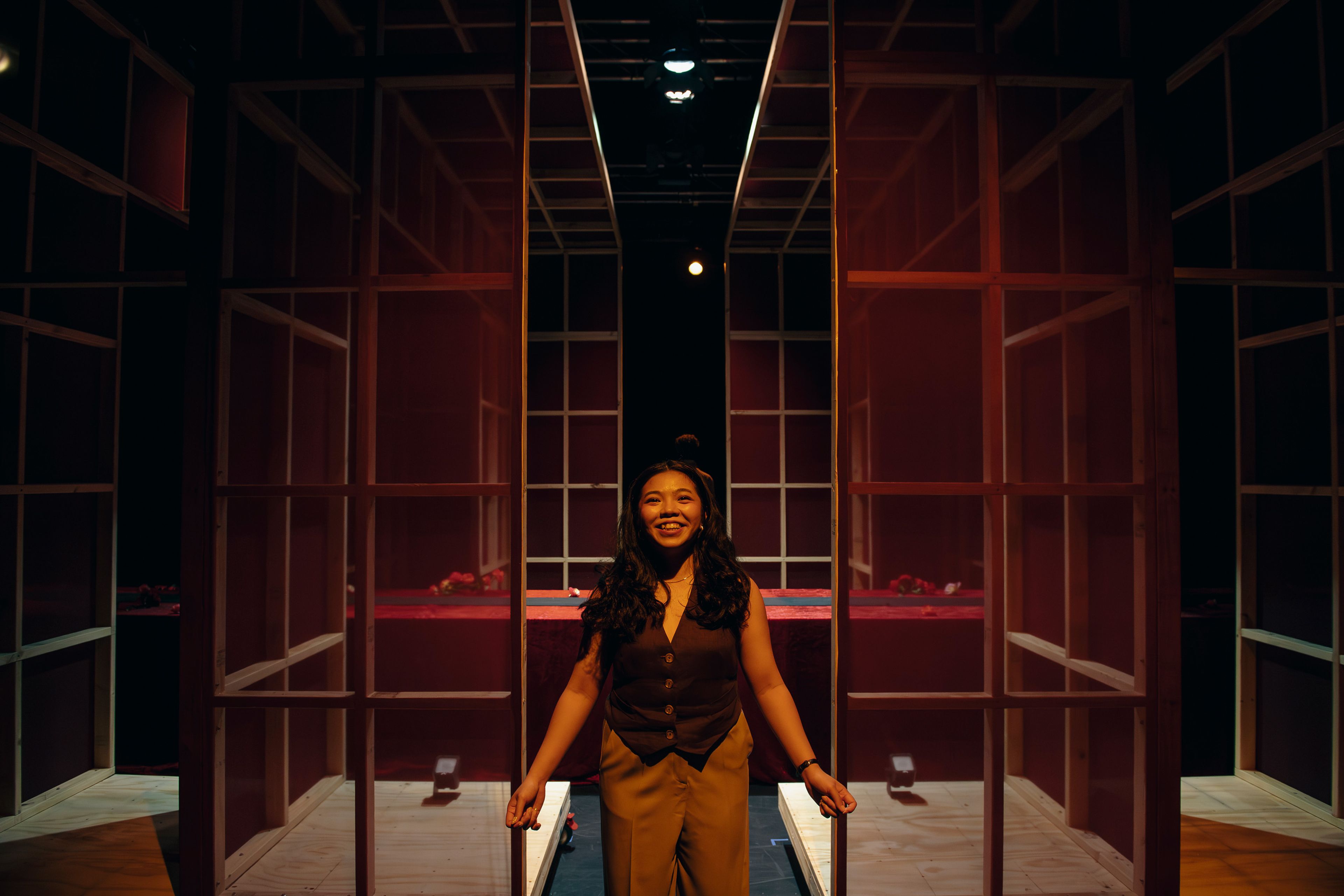 Theatre production photograph featuring a smiling actor standing in the centre of a wooden-framed structure, with soft red lighting creating a warm atmosphere.