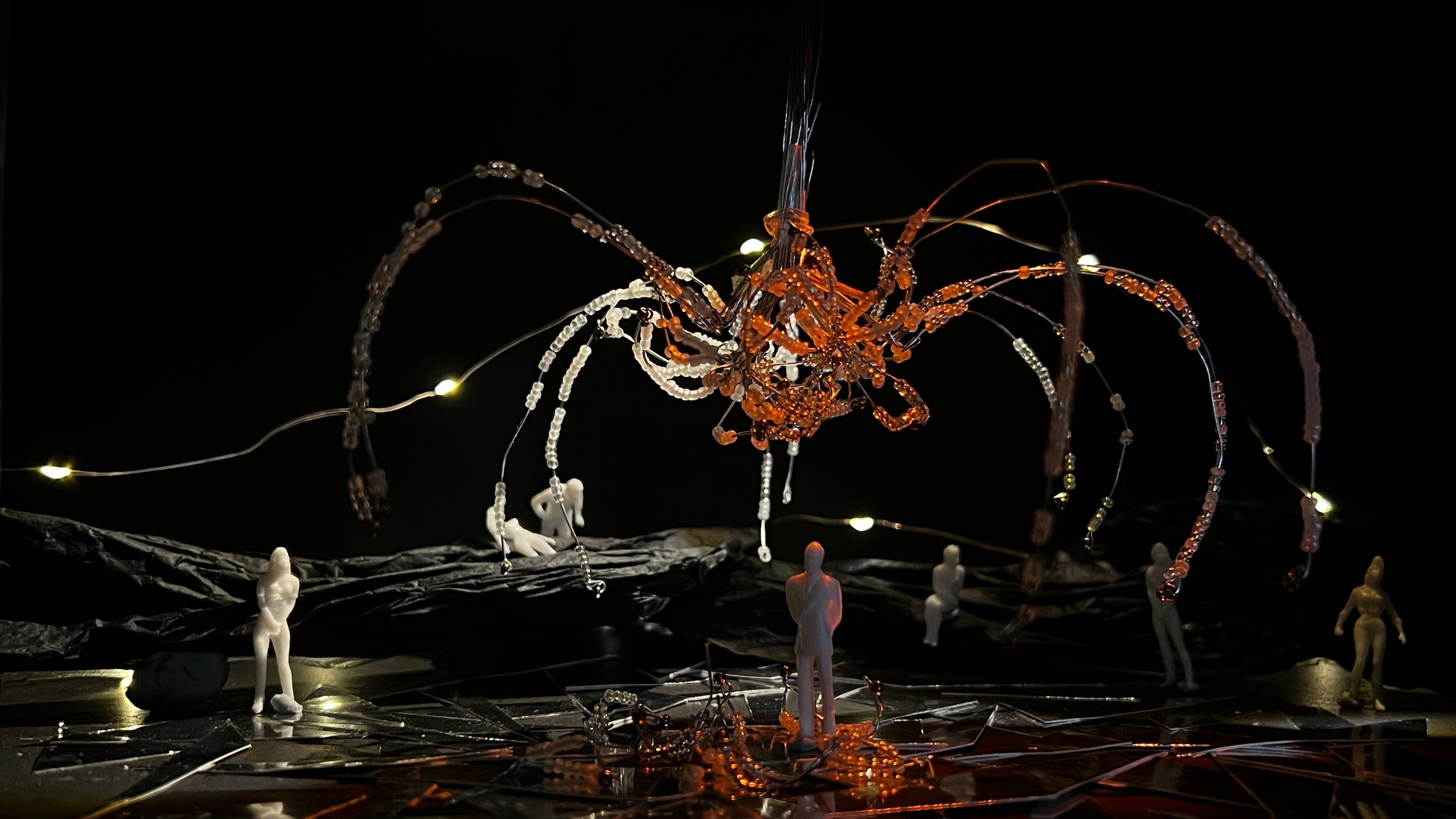 Miniature set design showing multiple white figures positioned beneath an amber-coloured beaded sculpture suspended overhead, with small lights scattered throughout the dark space