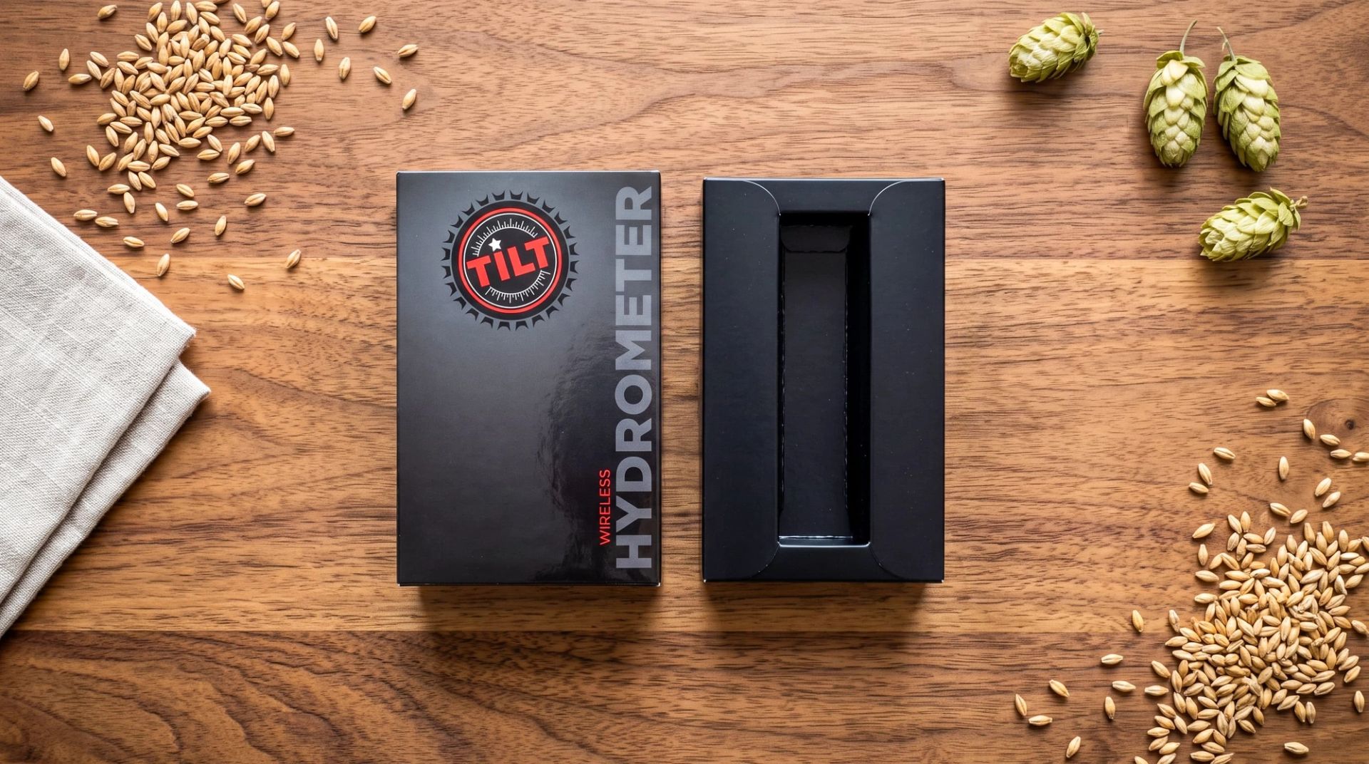 Closed TILT outer box sleeve next to an open inner tray with a protective slot, surrounded by hops and barley.