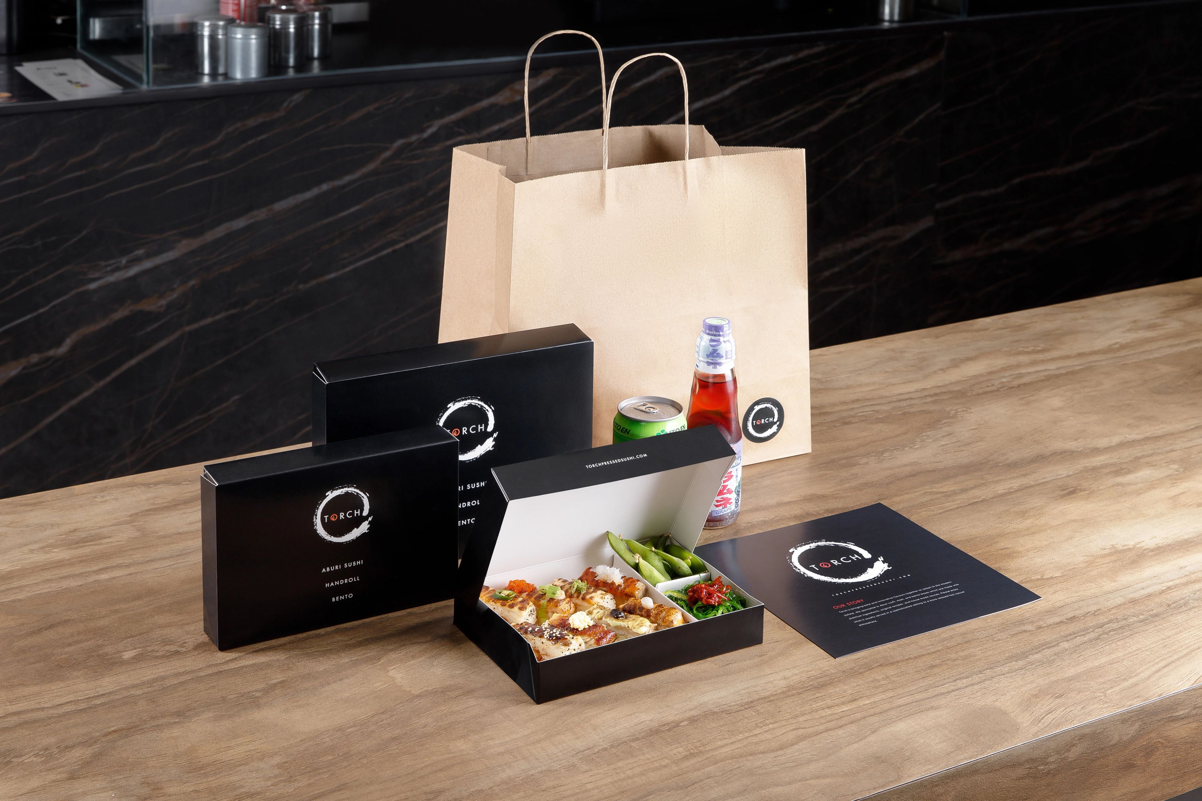A complete Torch Sushi takeout setup with an open sushi bento box, closed packaging, drinks, and a branded bag on a table.