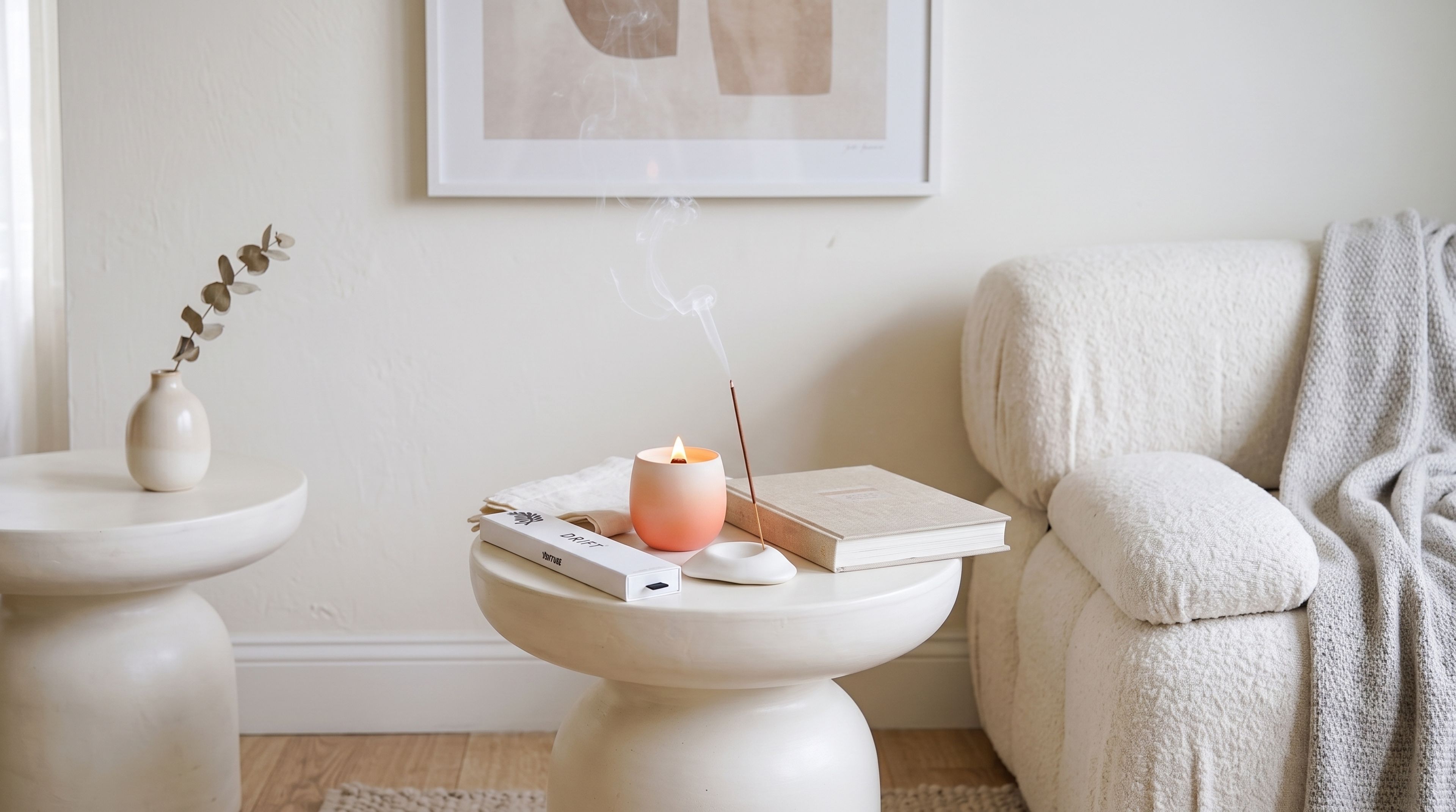 A lit peach candle, a smoking incense stick, and a Venture Drift incense box on a white pedestal table by a cream sofa.