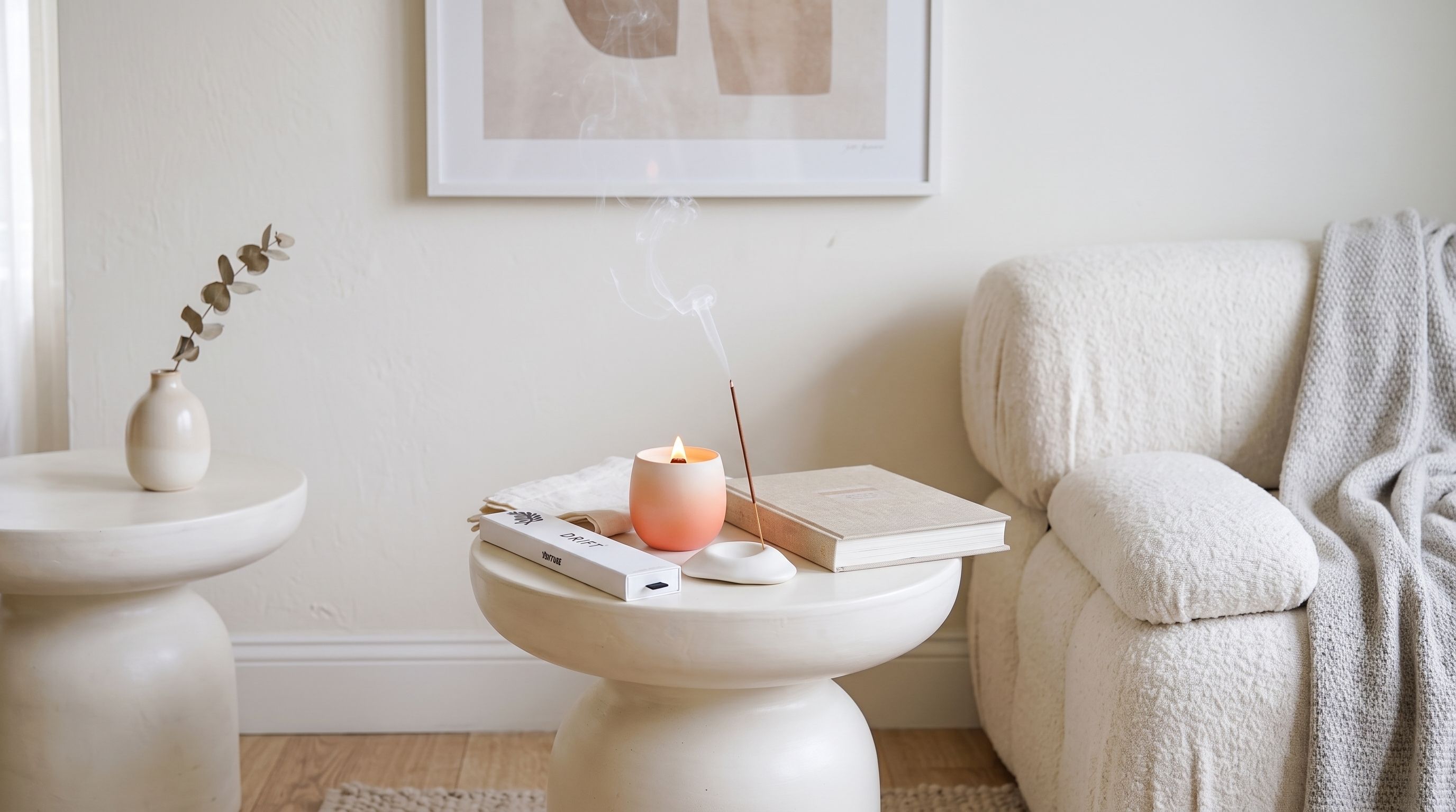 A lit peach candle, a smoking incense stick, and a Venture Drift incense box on a white pedestal table by a cream sofa.