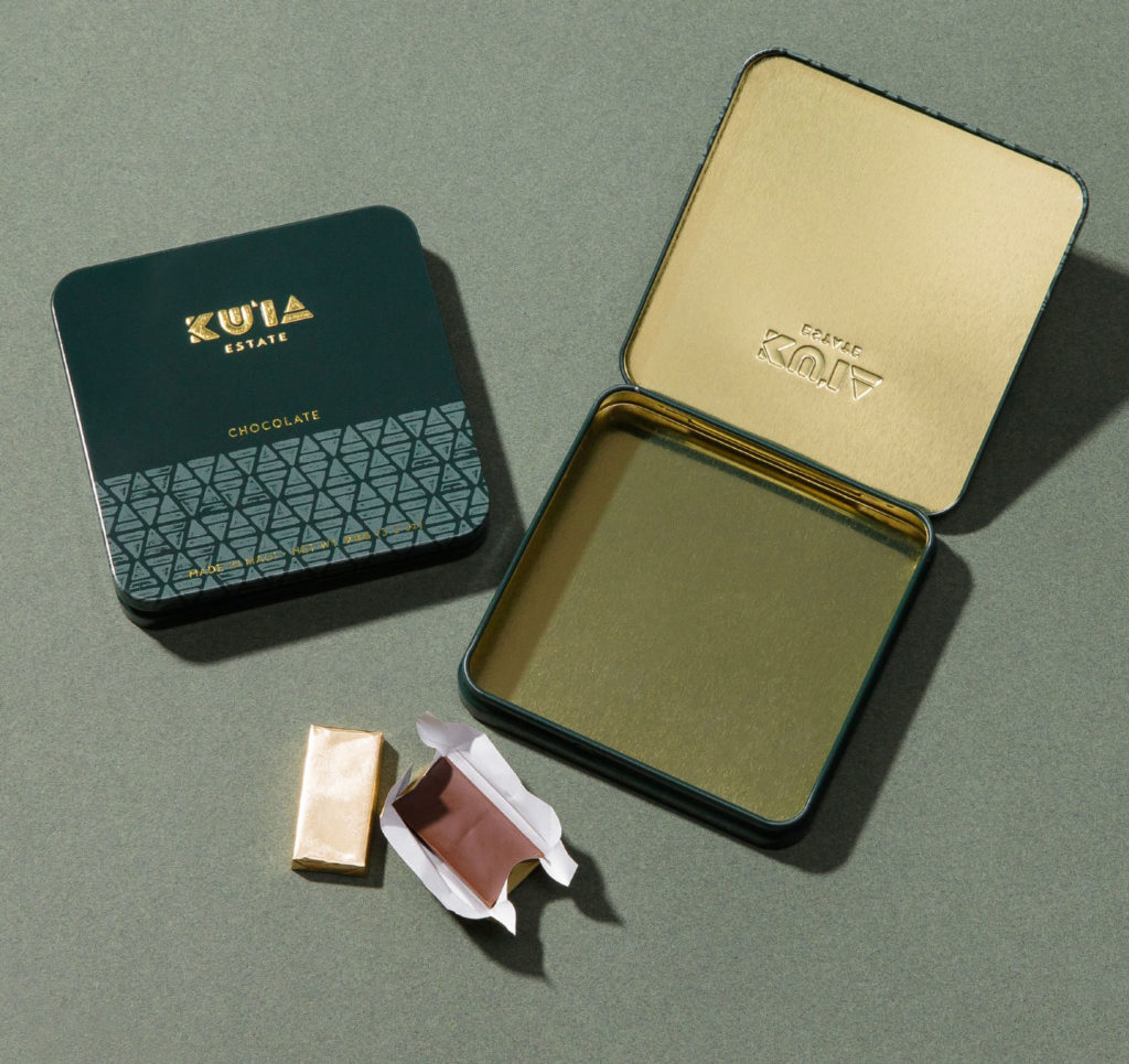 Top-down: a closed green Ku'ia Estate Chocolate tin, an open gold-lined tin, and two chocolates, foiled and bare, on green.