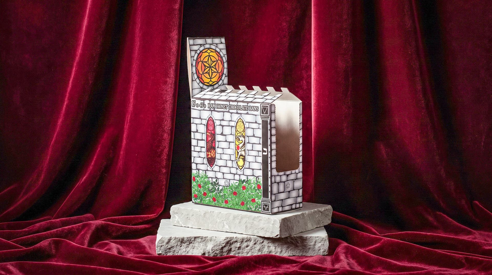 Side view of a castle-shaped chocolate display box with a side cutout, resting on stone slabs against red velvet.
