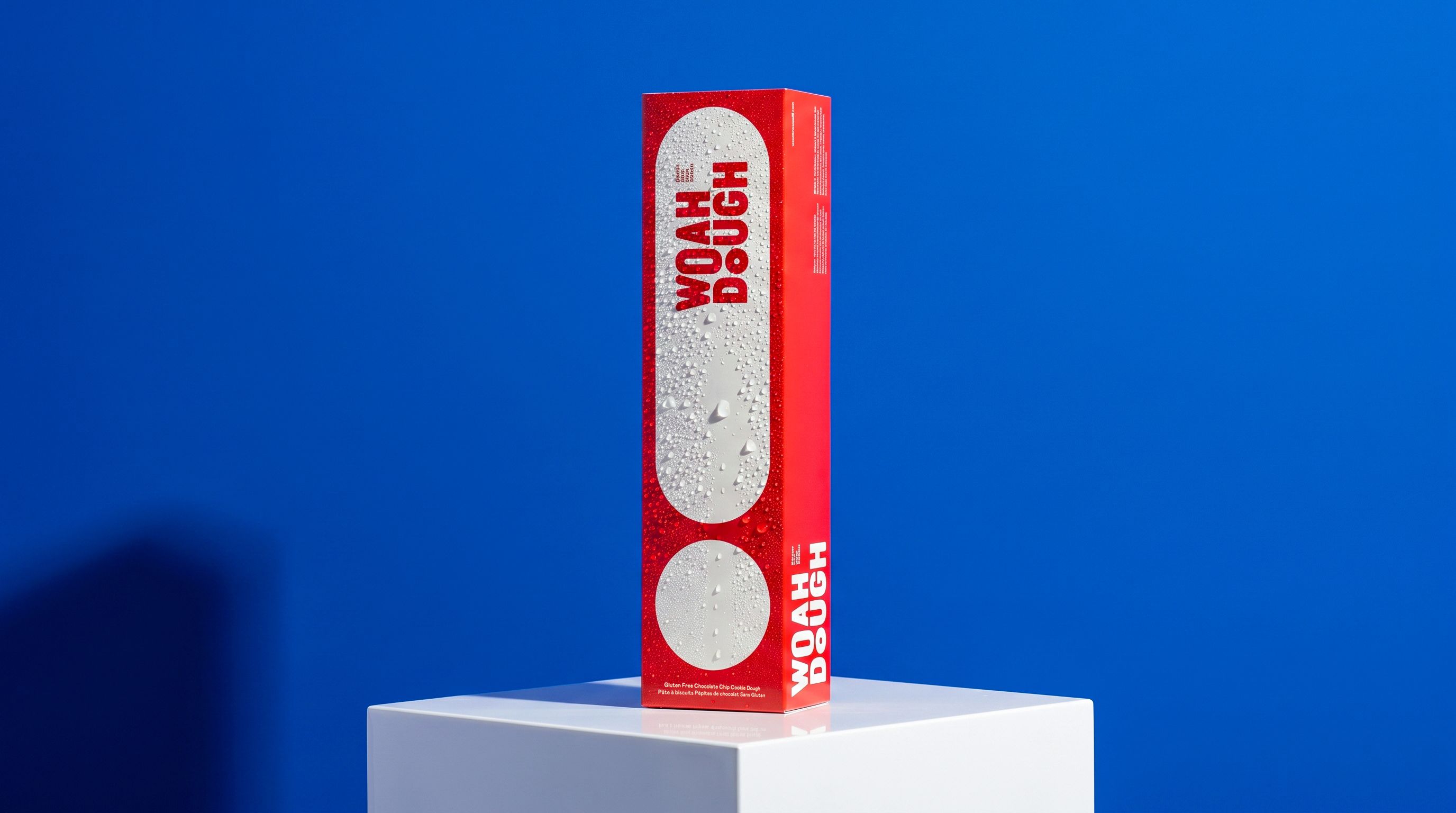 Tall red Woah Dough box with water condensation droplets, standing on a white pedestal against a bright blue background.