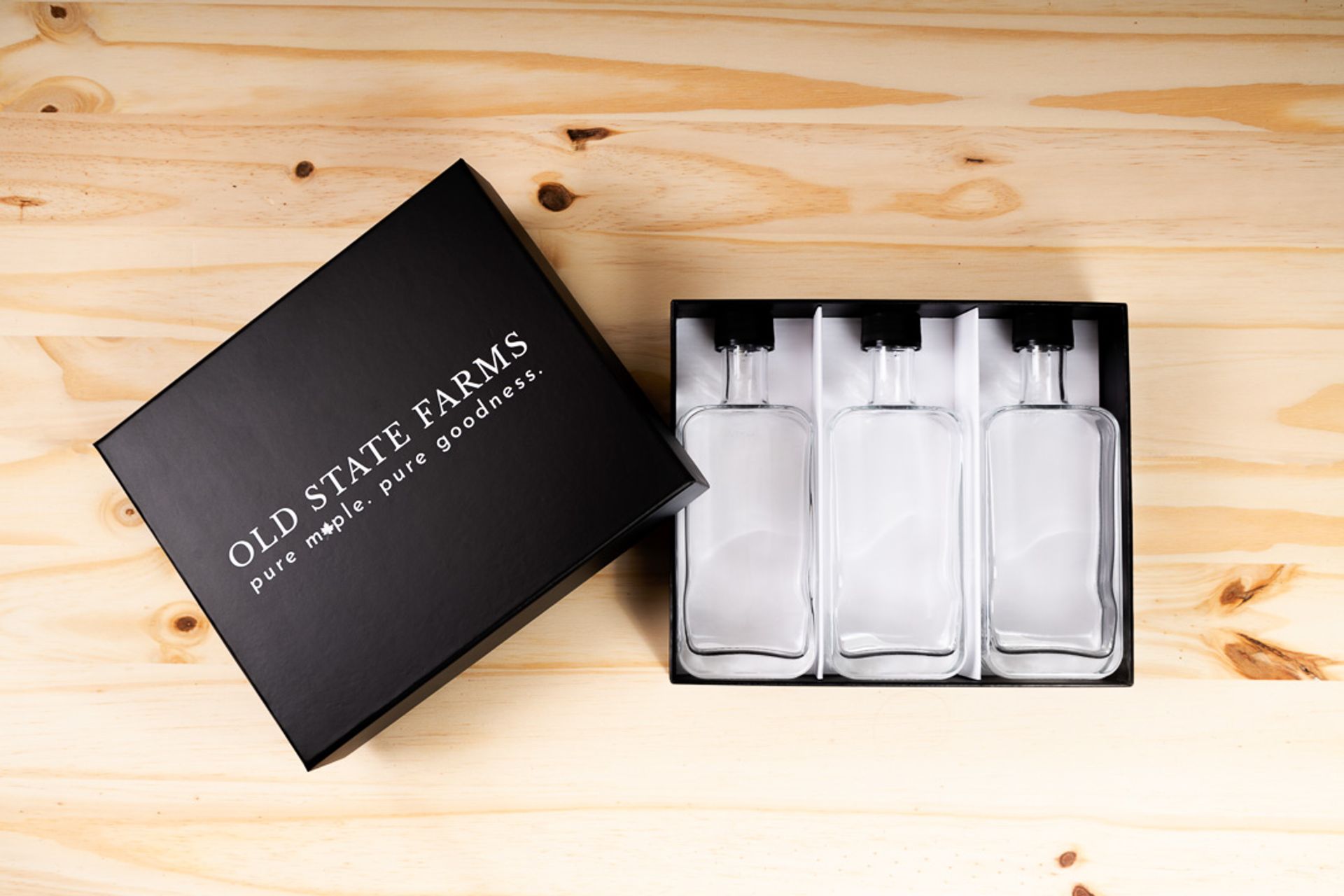 Top-down view of an open Old State Farms rigid box containing three empty glass syrup bottles separated by white dividers.