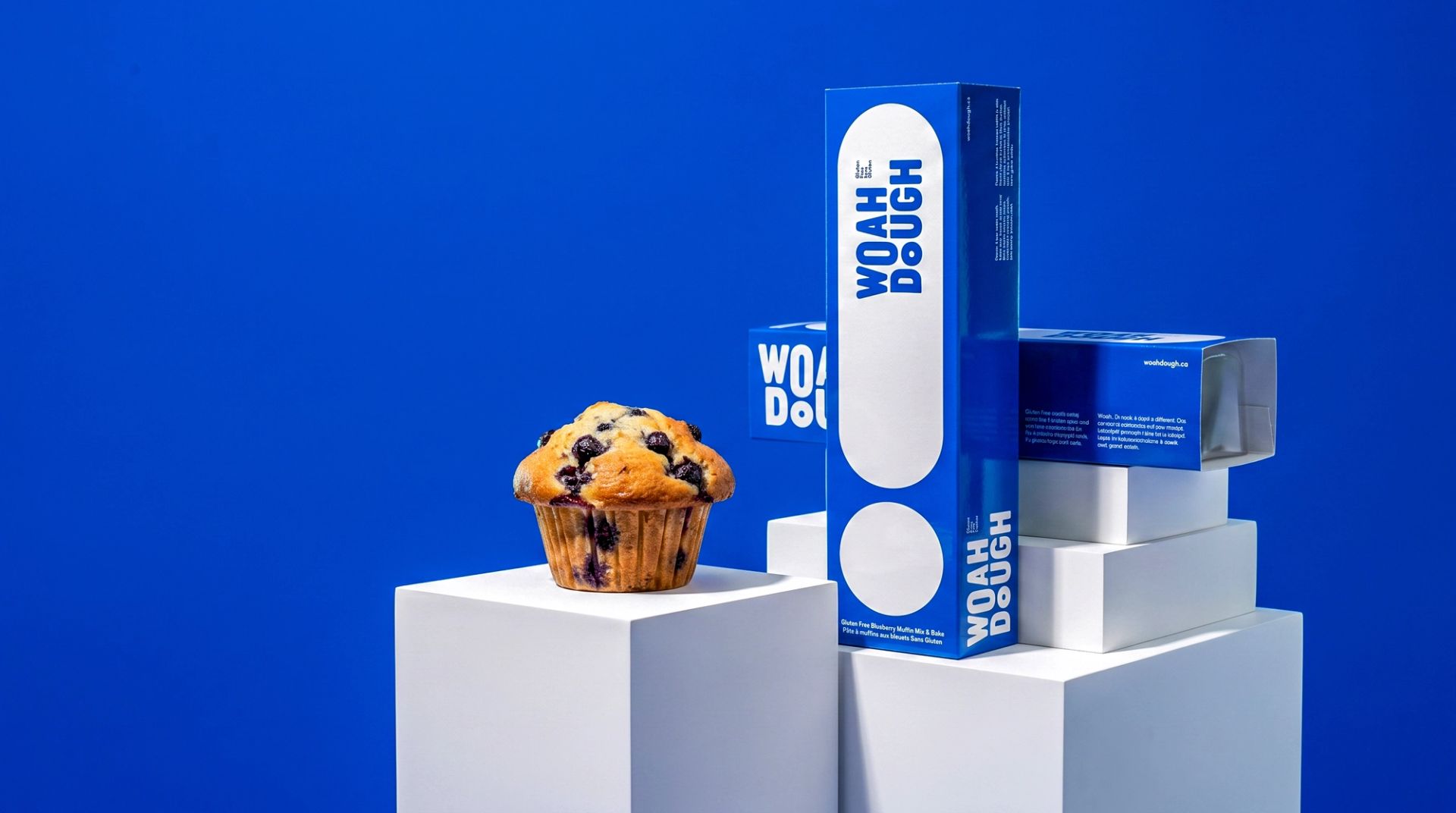 A blueberry muffin next to blue Woah Dough box on white pedestals against a vibrant blue background.
