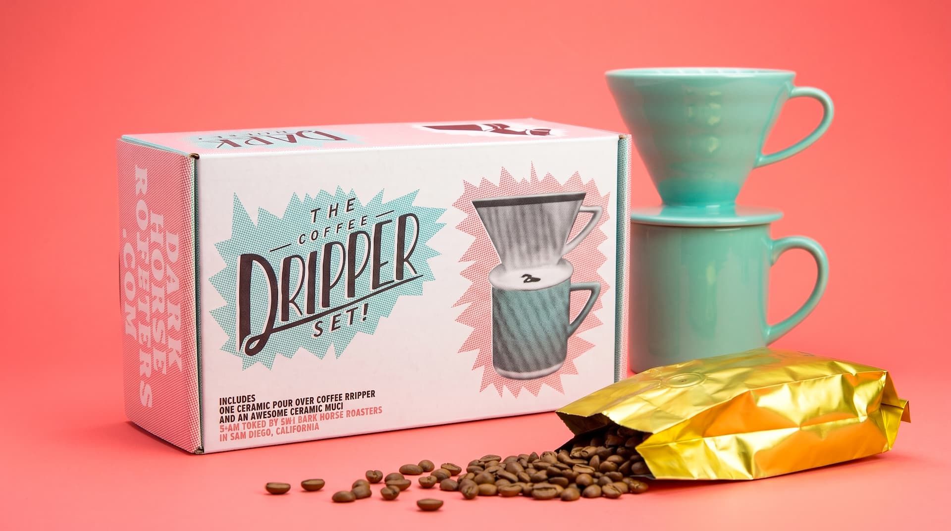 Closed Coffee Dripper Set box next to a stacked mint green mug and dripper, with a spilled gold bag of beans in front.