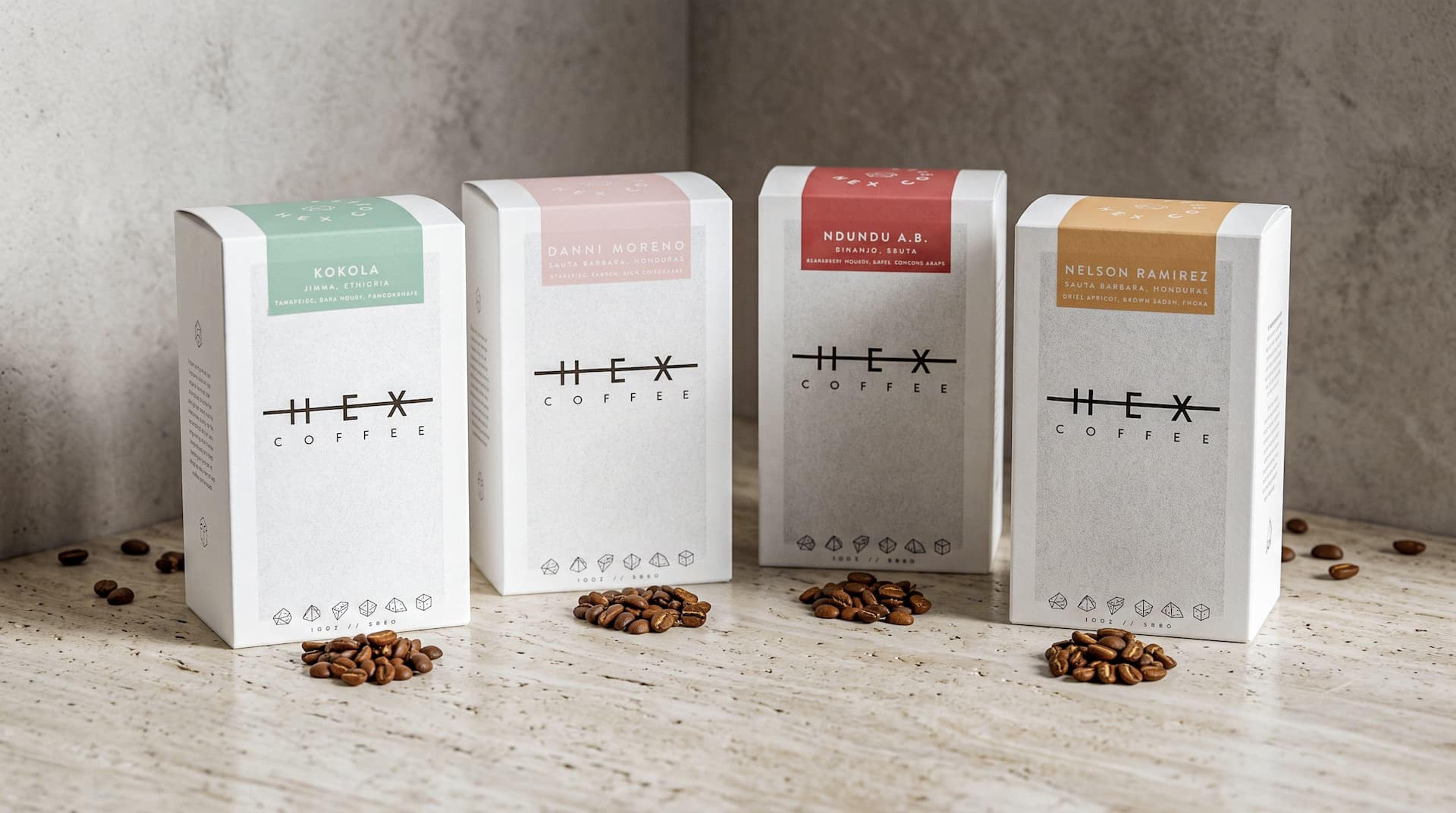 Four Hex Coffee boxes with distinct colored labels standing in a row, each with a small pile of coffee beans in front.