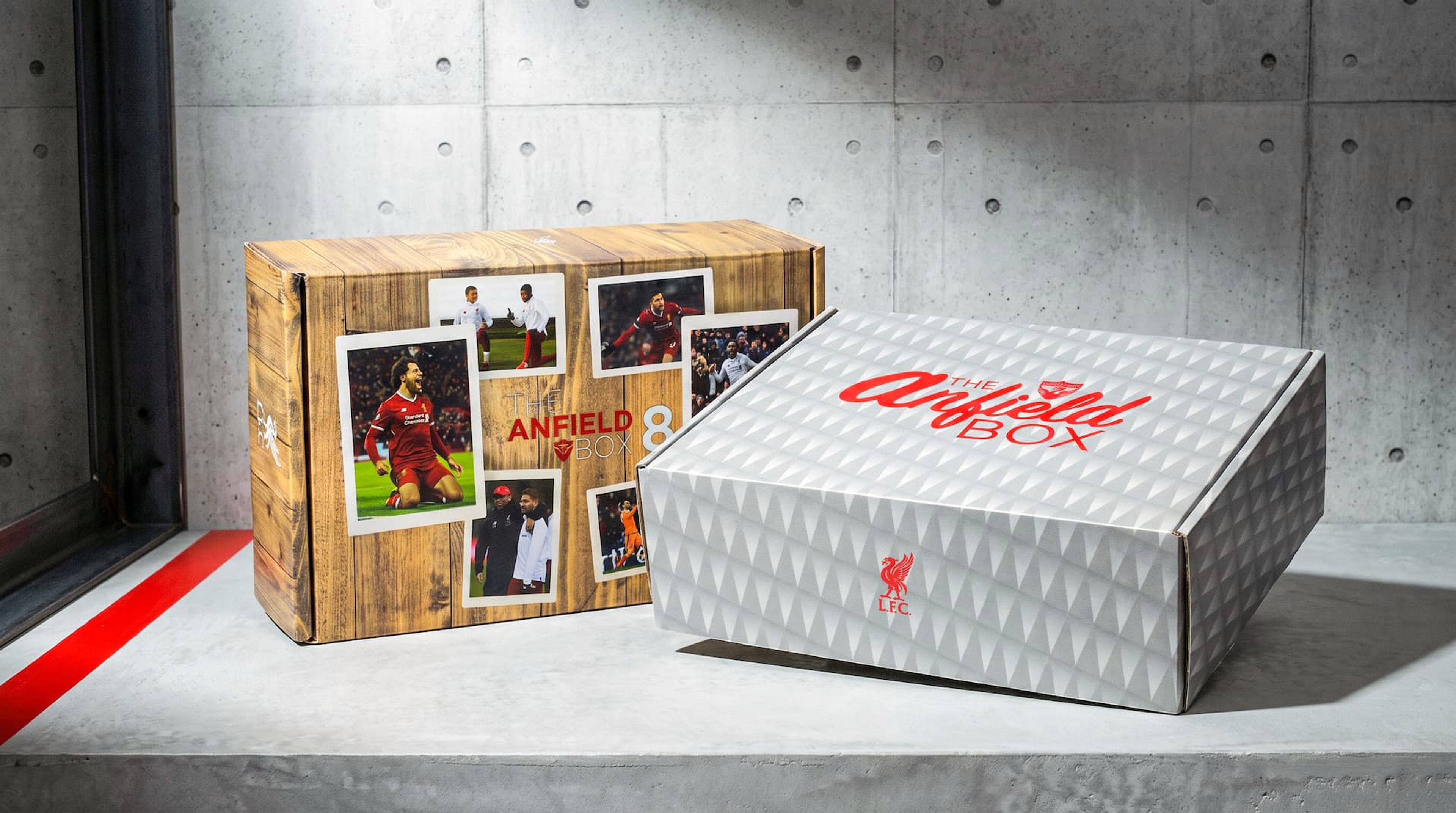 Two Anfield Box packages on concrete: one with a wood-grain design and player photos, the other white with a geometric pattern.