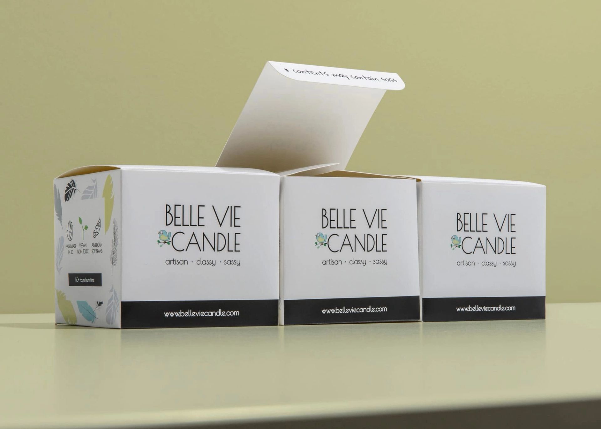 Three white Belle Vie Candle boxes lined up; the center box's open lid reveals the text "contents may contain sass".