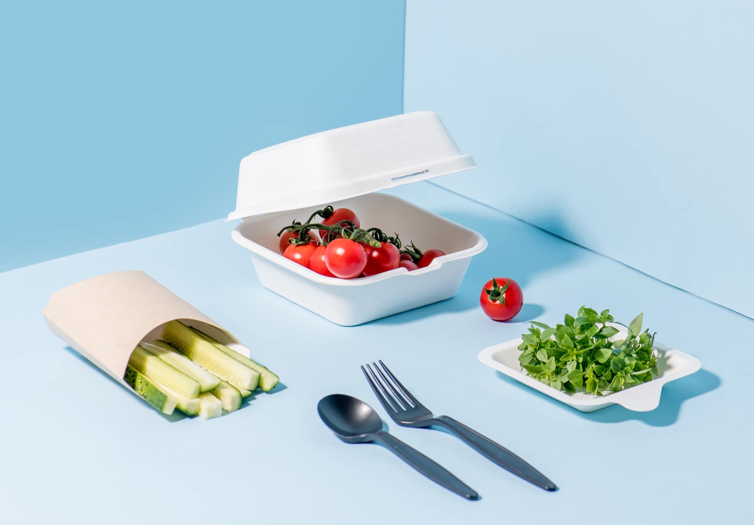 White and brown bioplastic cornstarch packaging container with food and utensils on a blue background.