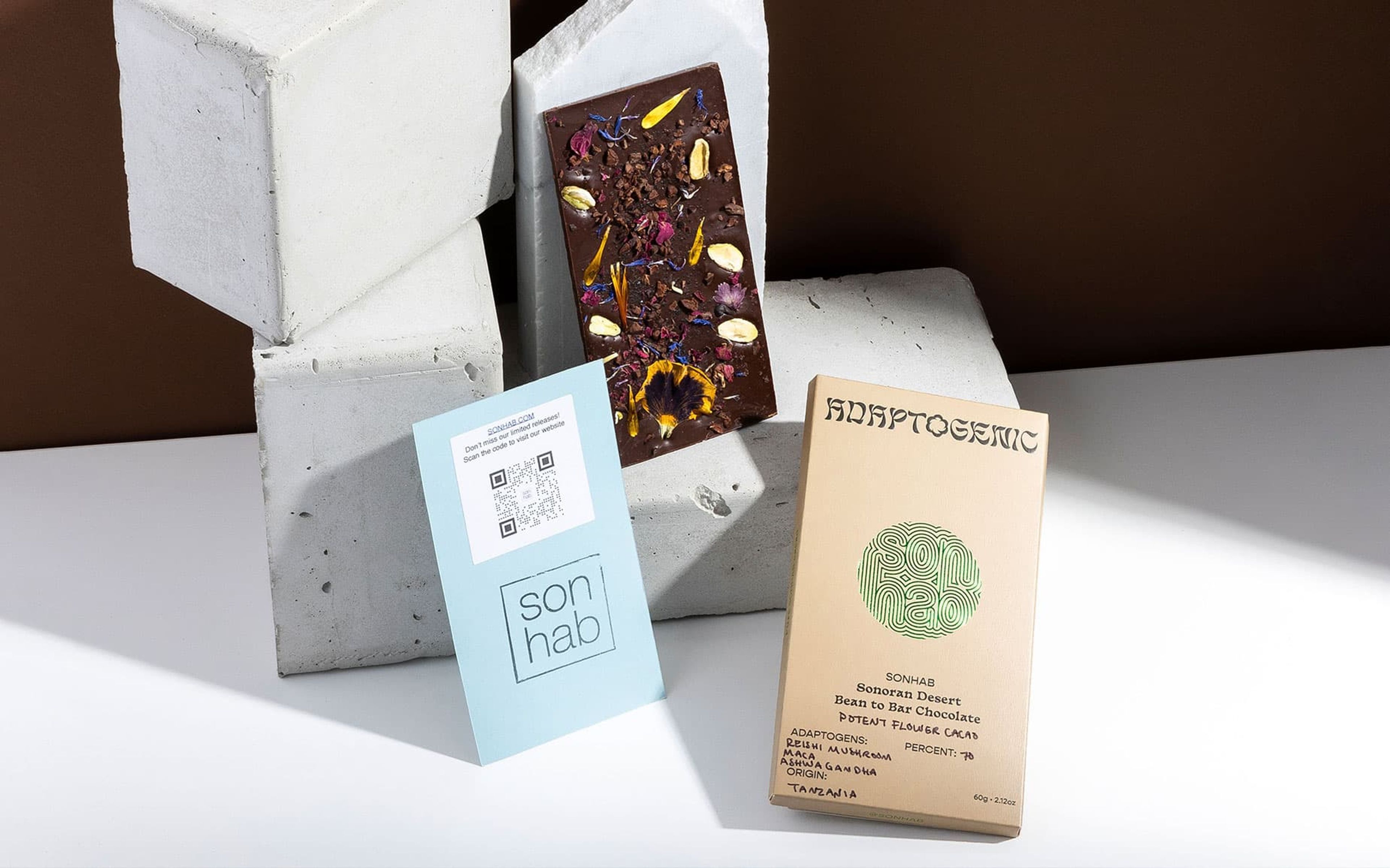 Sonhab Adaptogenic chocolate box, floral chocolate bar, and blue QR code card displayed on grey concrete blocks.