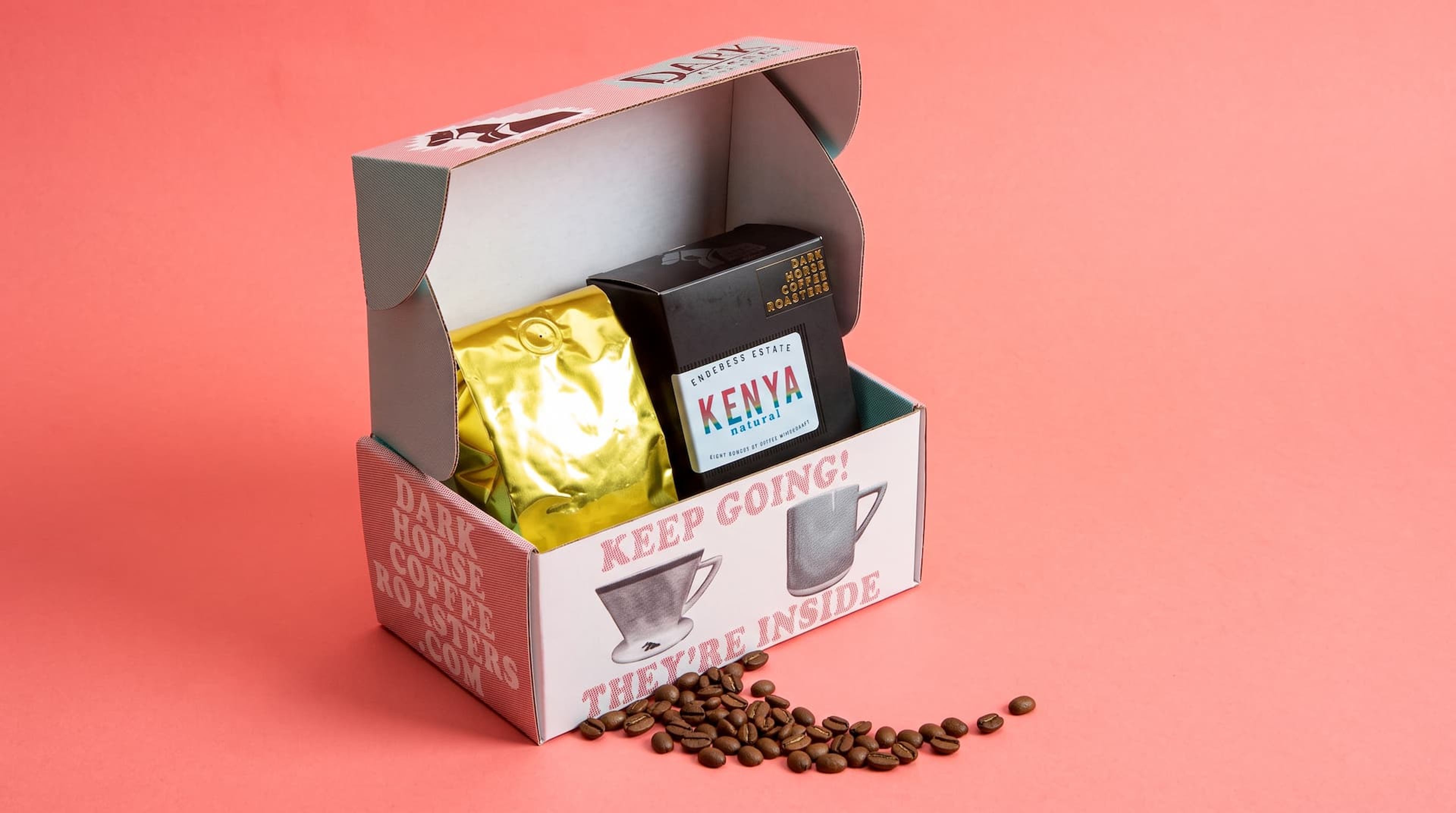 Open Dark Horse coffee box containing a gold foil bag and a black Kenya Natural box, with loose beans on a pink background.