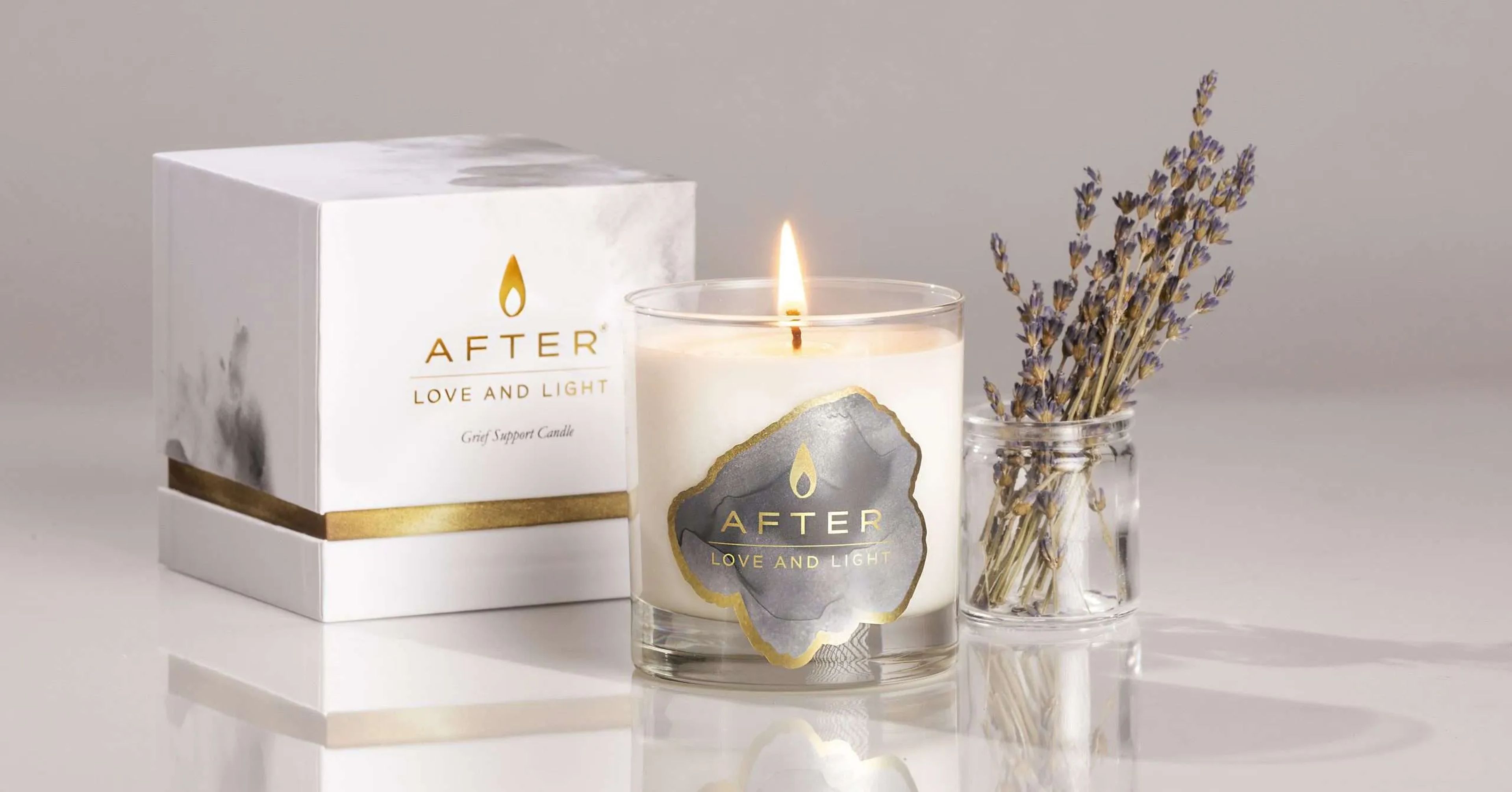 A lit AFTER Love and Light candle sitting next to its closed white, gold-rimmed rigid box on a marble surface.