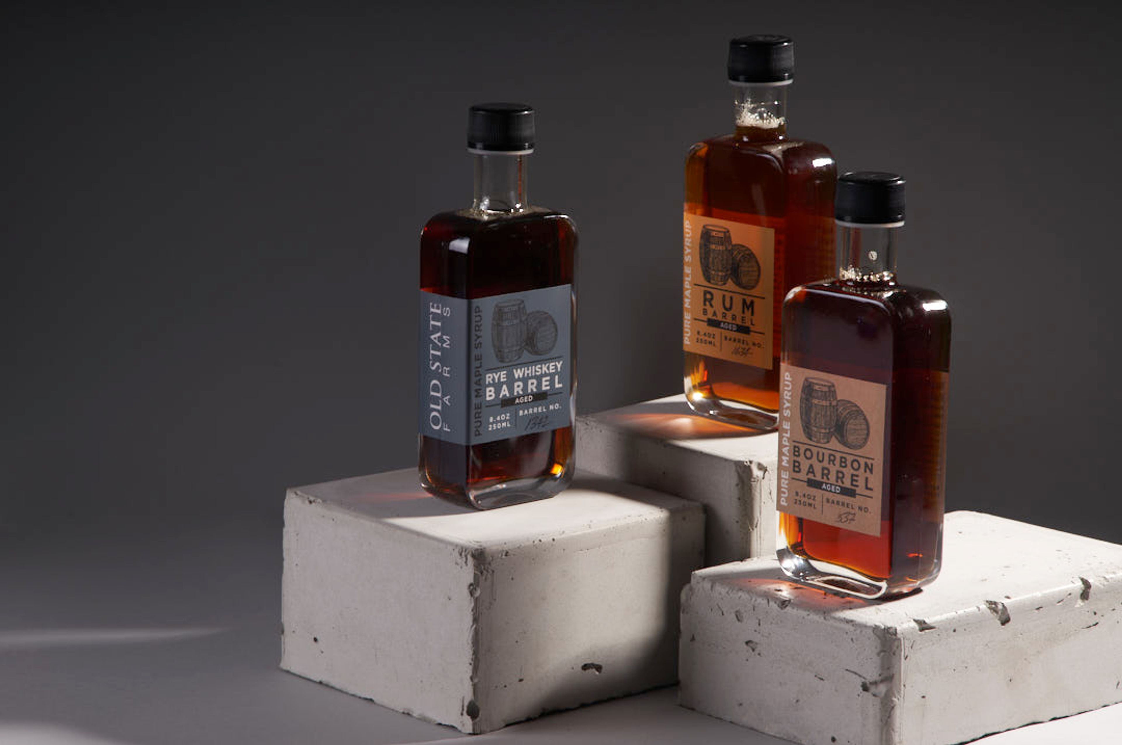 Three bottles of Old State Farms barrel-aged maple syrup—Rye, Rum, and Bourbon—displayed on concrete blocks.