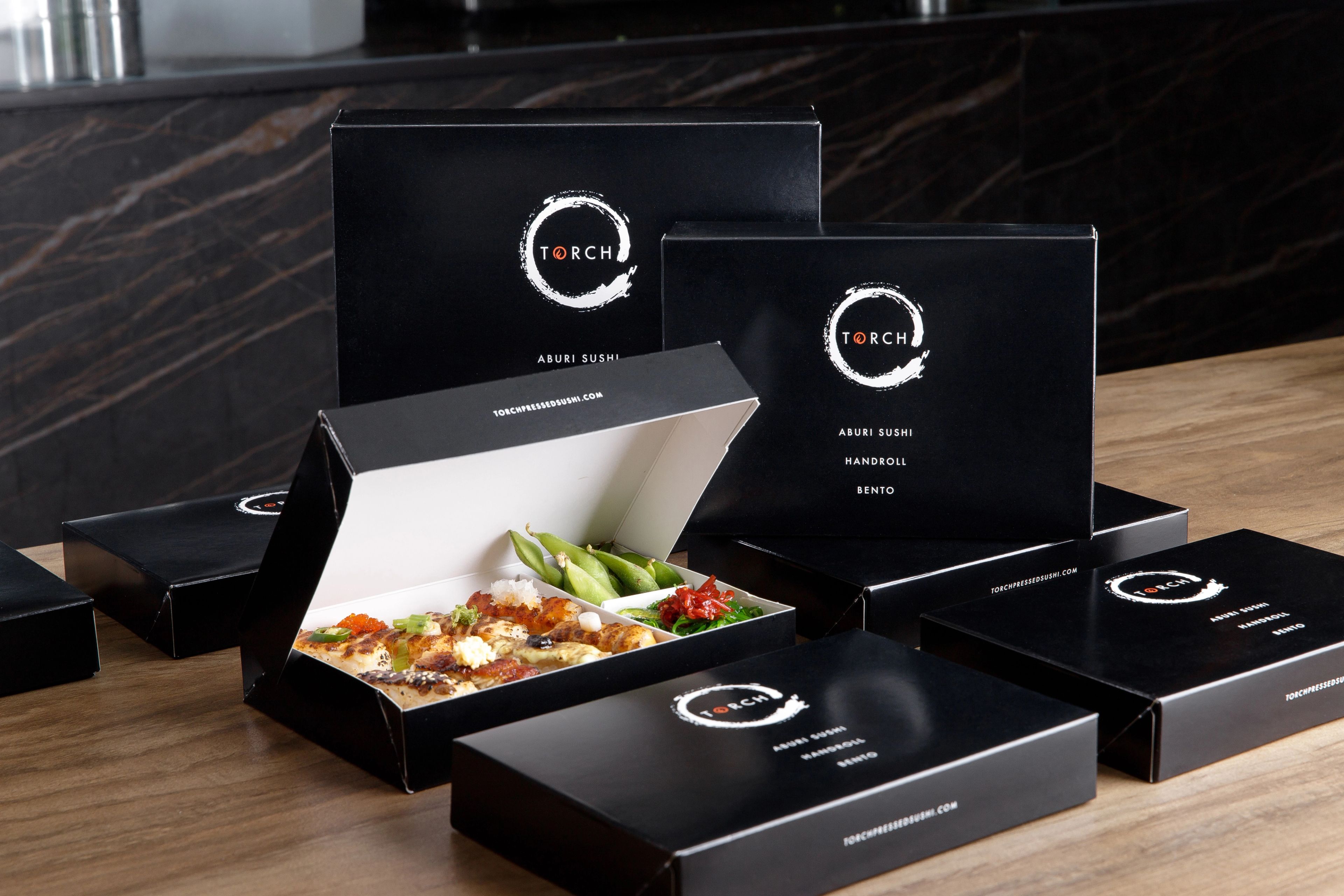 Open Torch Sushi bento box with seared sushi and edamame in front of stacked closed black packaging on a wooden table.
