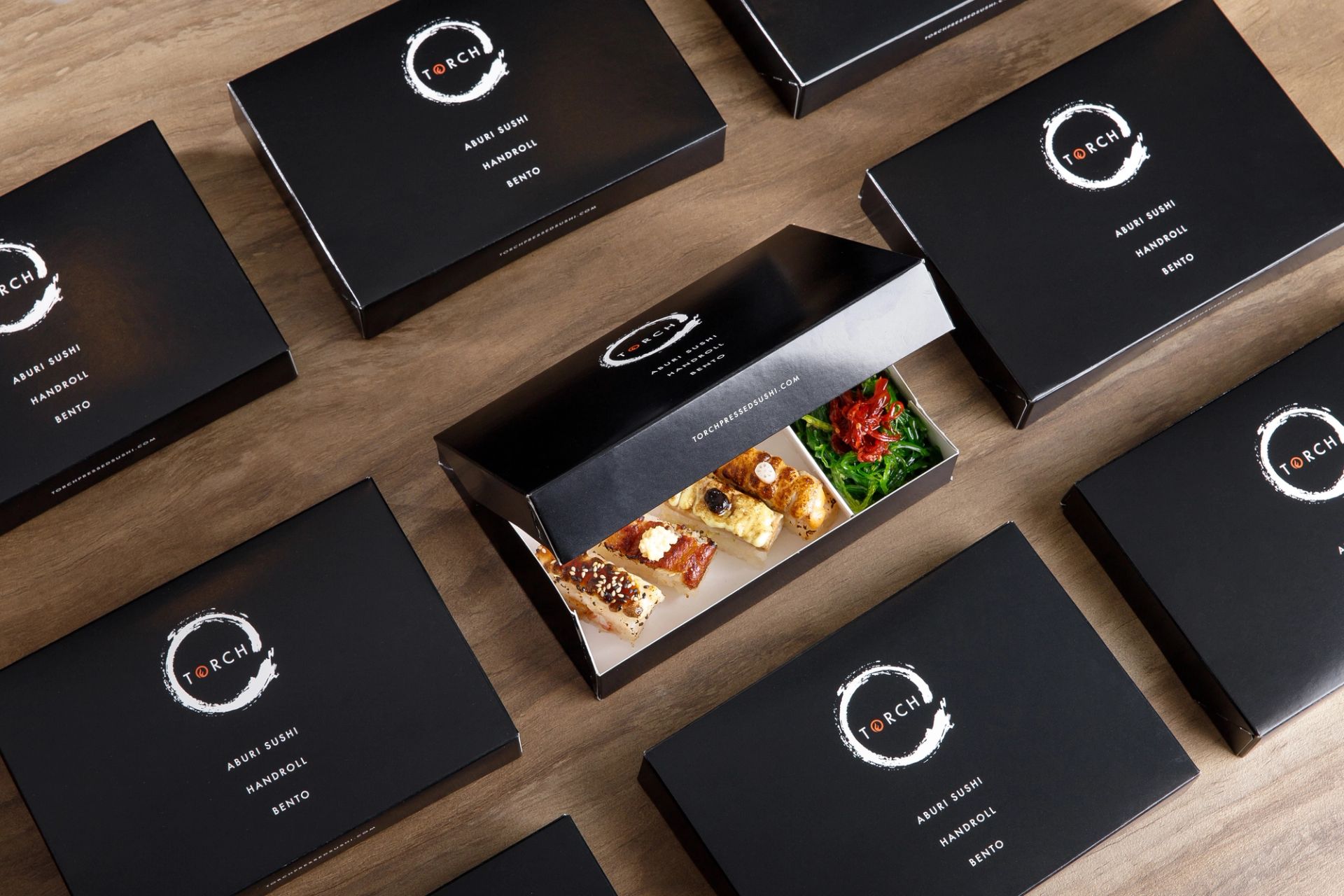 Grid of closed black Torch bento boxes on a table, with one open box in the center displaying aburi sushi and salad.