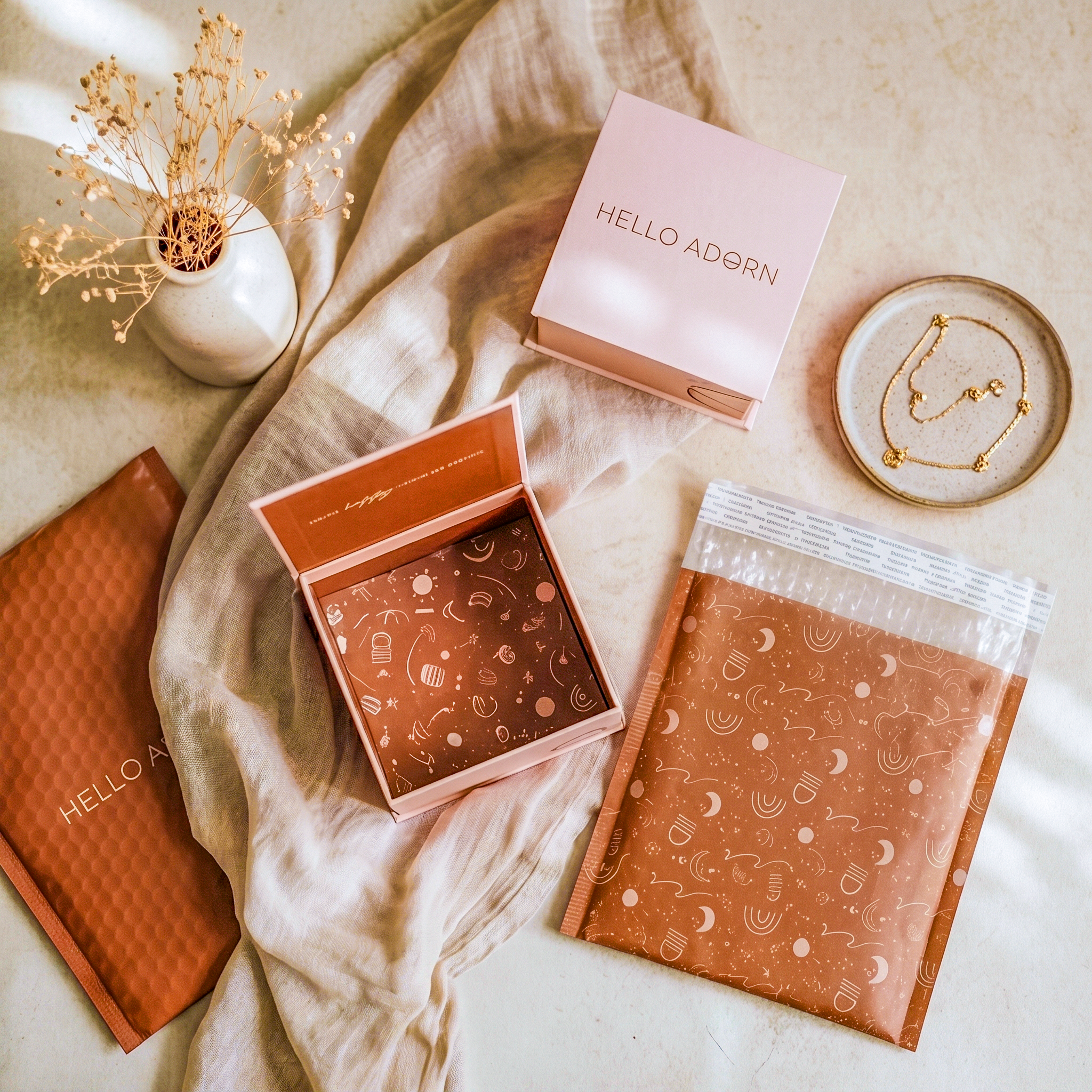 Flat lay of Hello Adorn jewelry packaging on beige fabric: pink boxes, rust mailers, a gold necklace, and dried flowers.