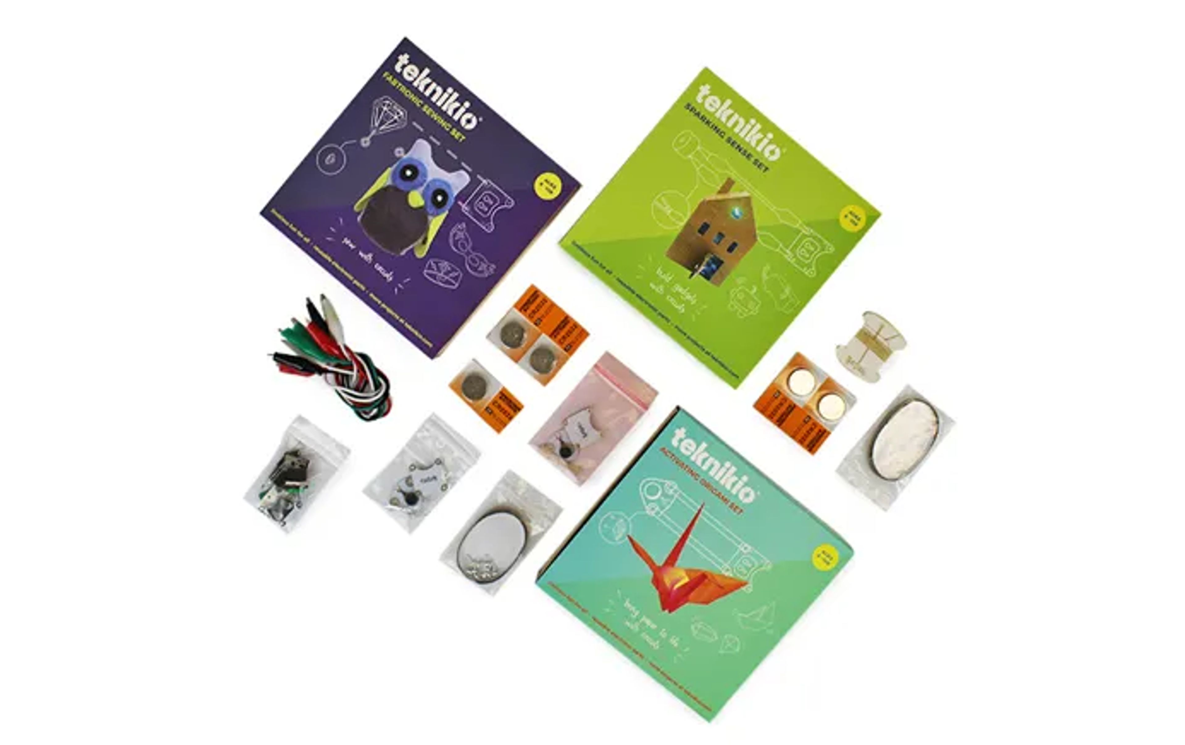 Three colorful Teknikio boxes surrounded by scattered electronic craft components like batteries, clips, and LEDs.