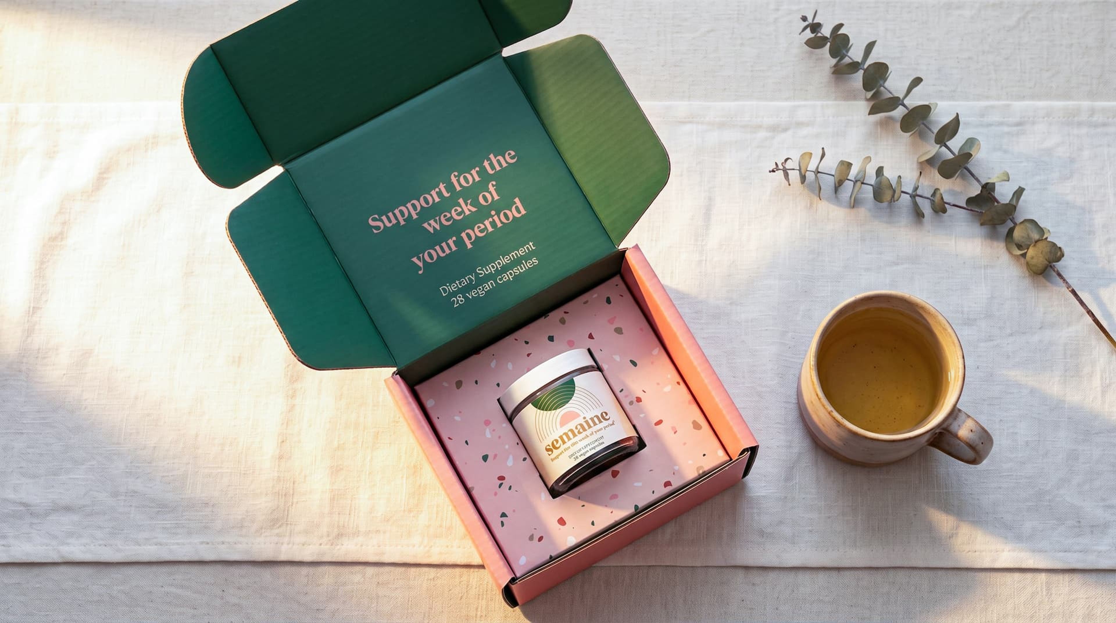 Top-down view of an open Semaine box holding a supplement jar, next to a mug of tea and a eucalyptus sprig.