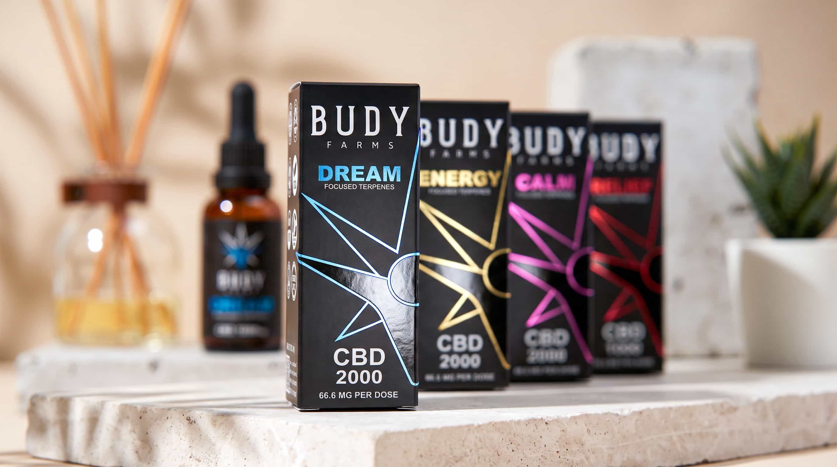 Four black Budy Farms CBD boxes—Dream, Energy, Calm, Relief—with a dropper bottle blurred in the background.