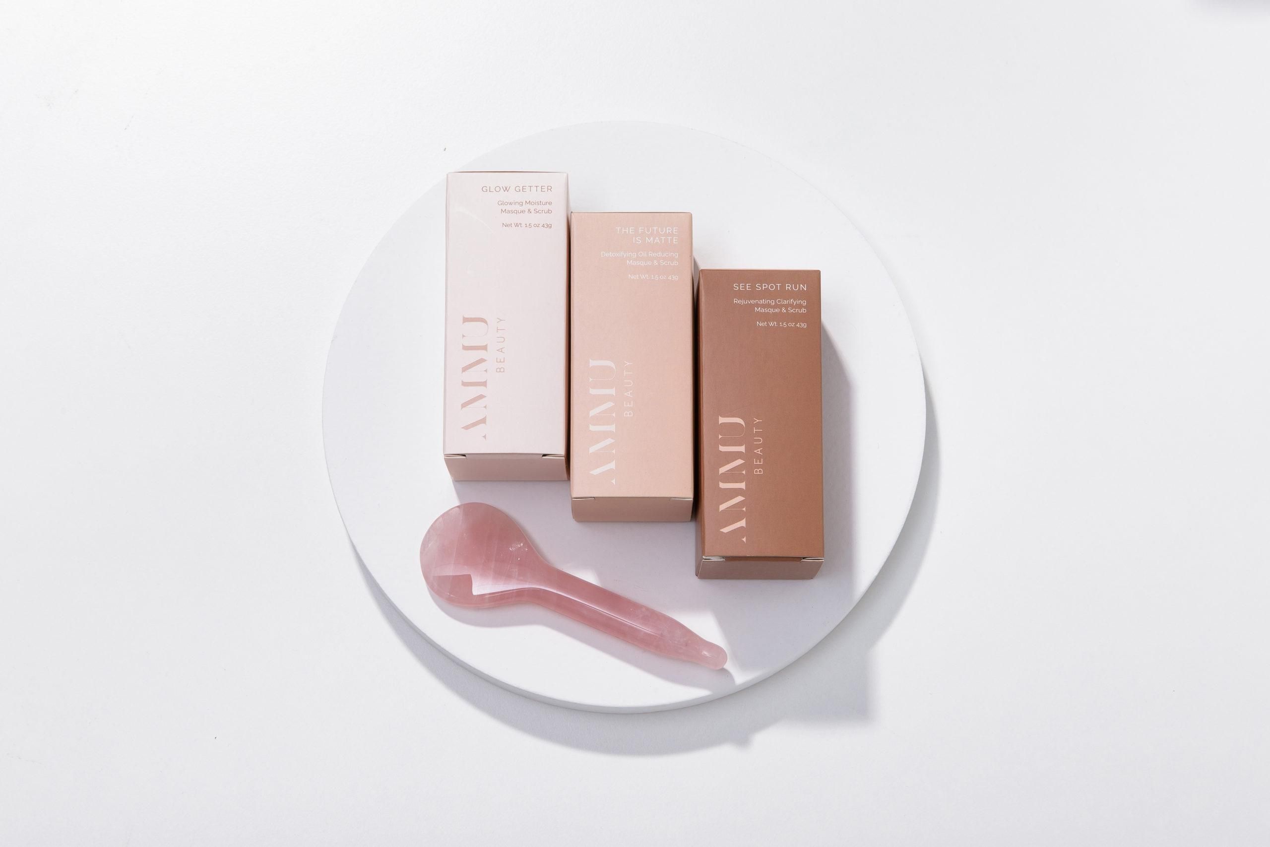 Three Ammu Beauty masque boxes and a rose quartz applicator spoon on a round white tray.