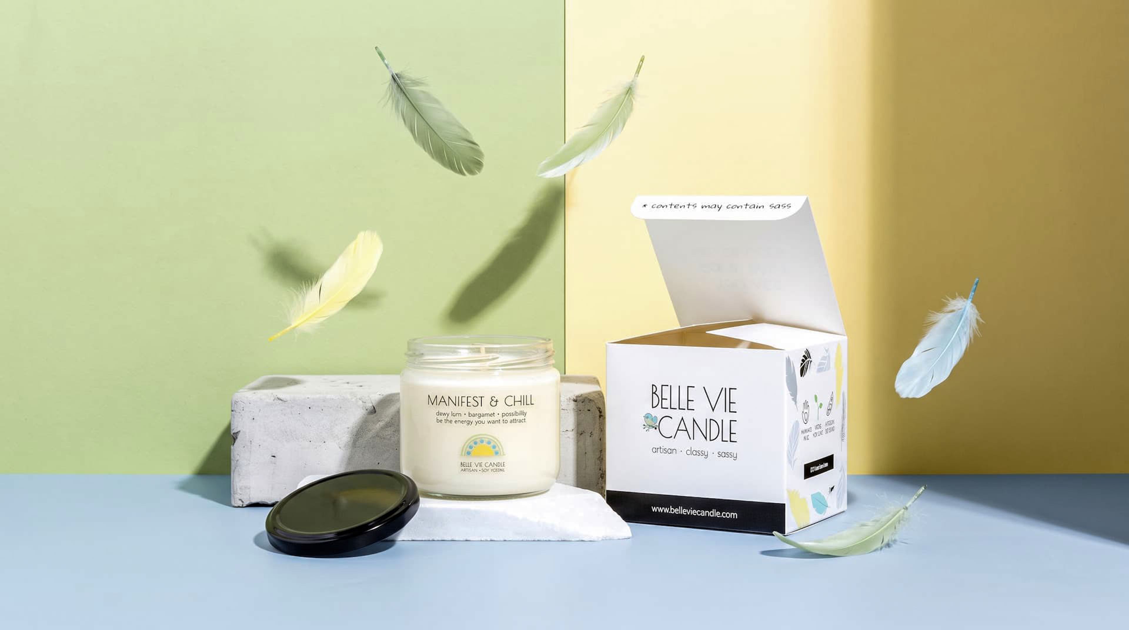 Open Belle Vie Candle jar and printed box on concrete blocks with pastel feathers floating against a green and yellow wall.
