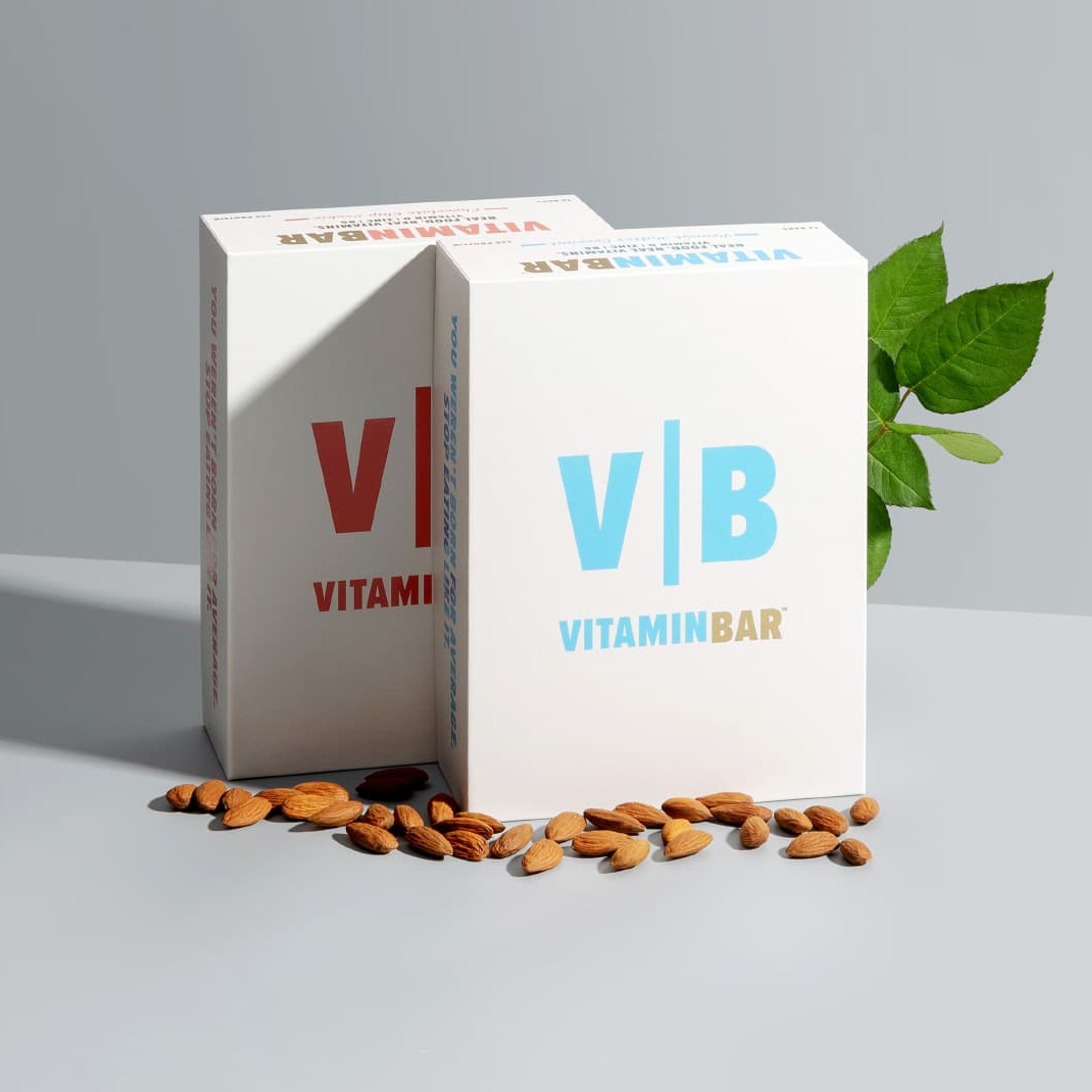 Two upright Vitamin Bar boxes in red and blue standing on a grey surface with scattered almonds and green leaves.