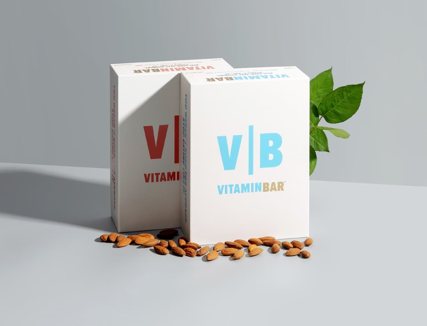 Two upright Vitamin Bar boxes in red and blue standing on a grey surface with scattered almonds and green leaves.