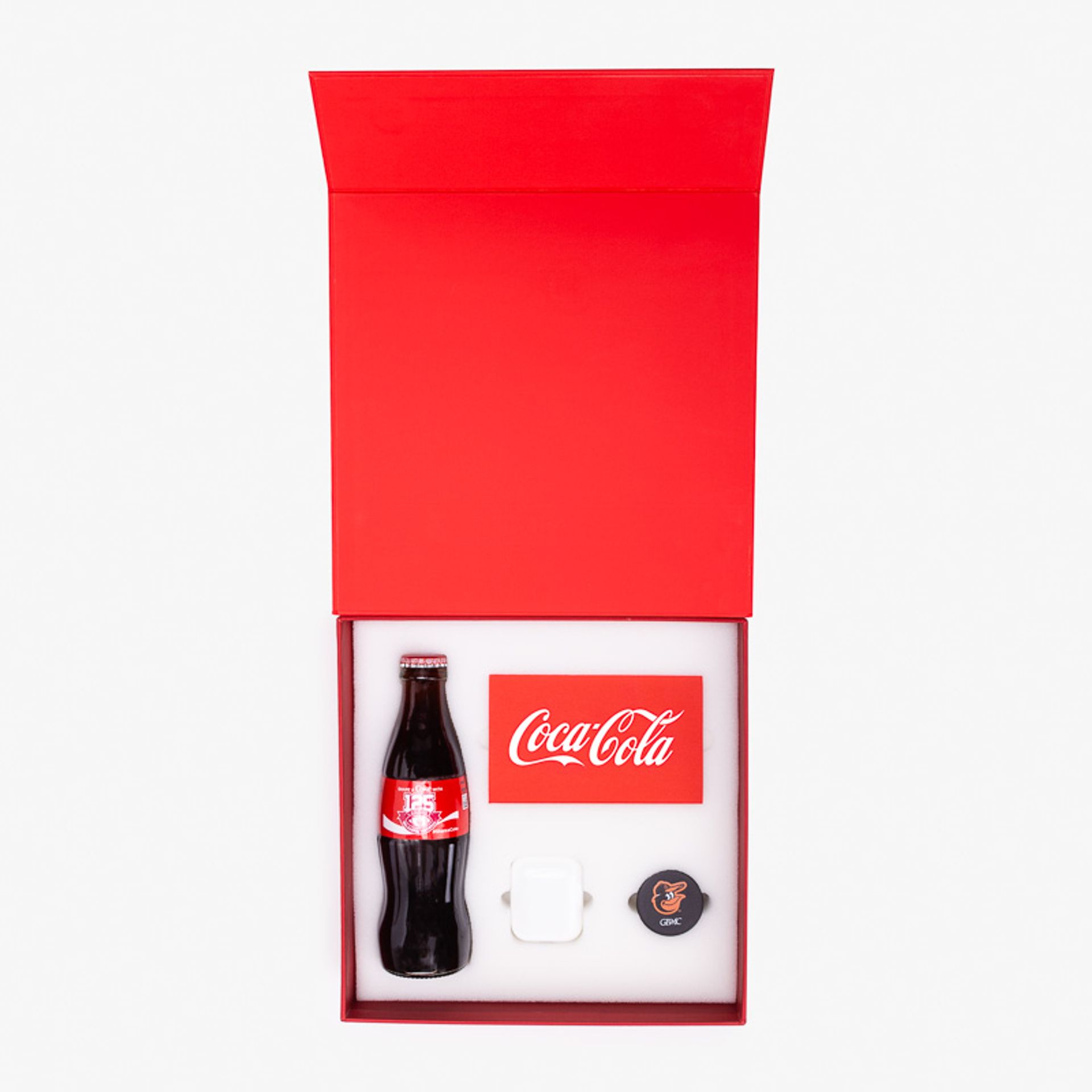 Top view of an open Coca-Cola gift box displaying a glass bottle, branded card, AirPods, and PopSocket.