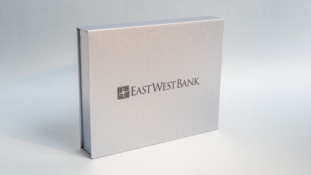 A closed, rectangular metallic silver presentation box standing upright, featuring the East West Bank logo on the cover.