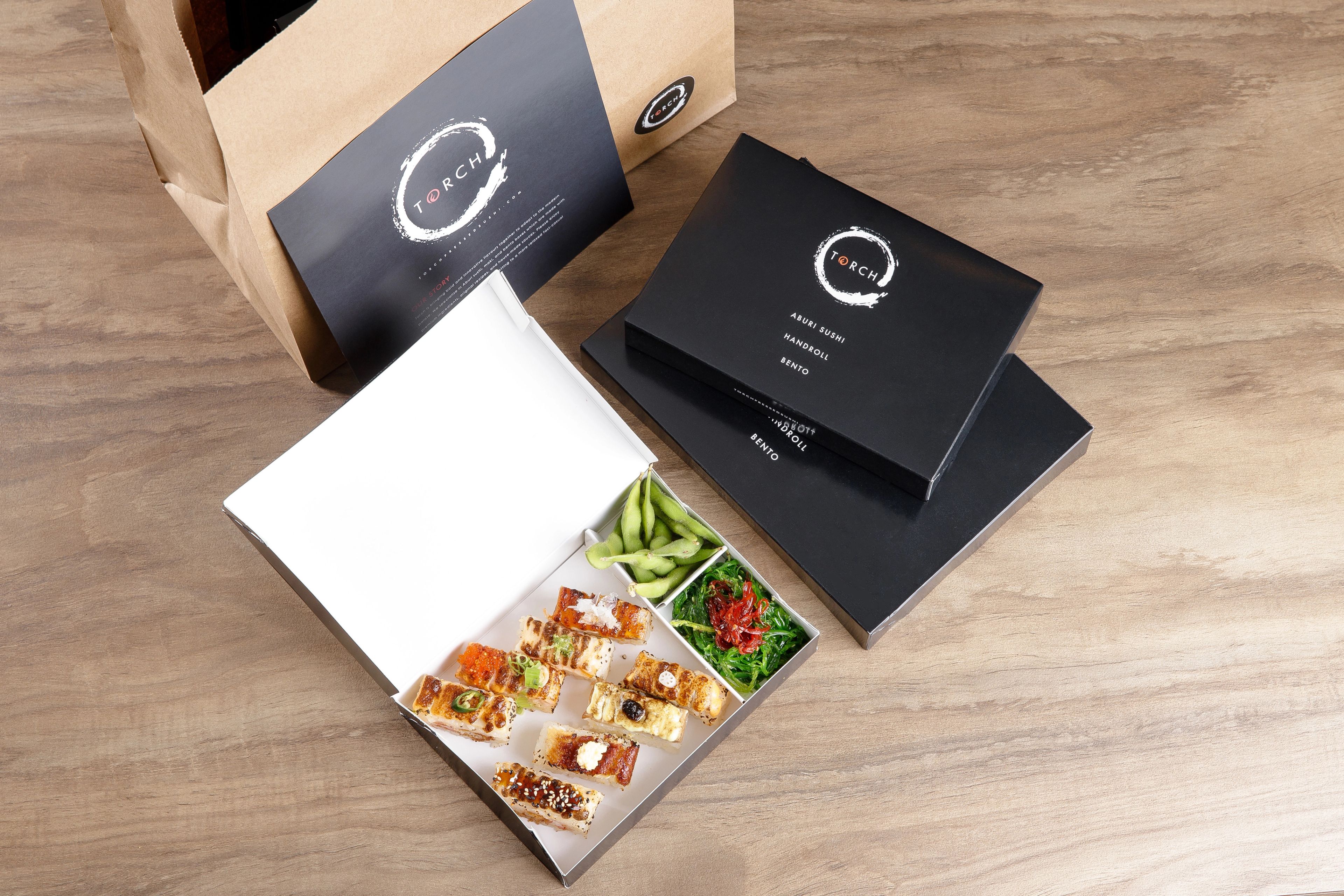 Open Torch Sushi bento box with aburi sushi, edamame, and salad next to closed boxes and a takeout bag on a table.