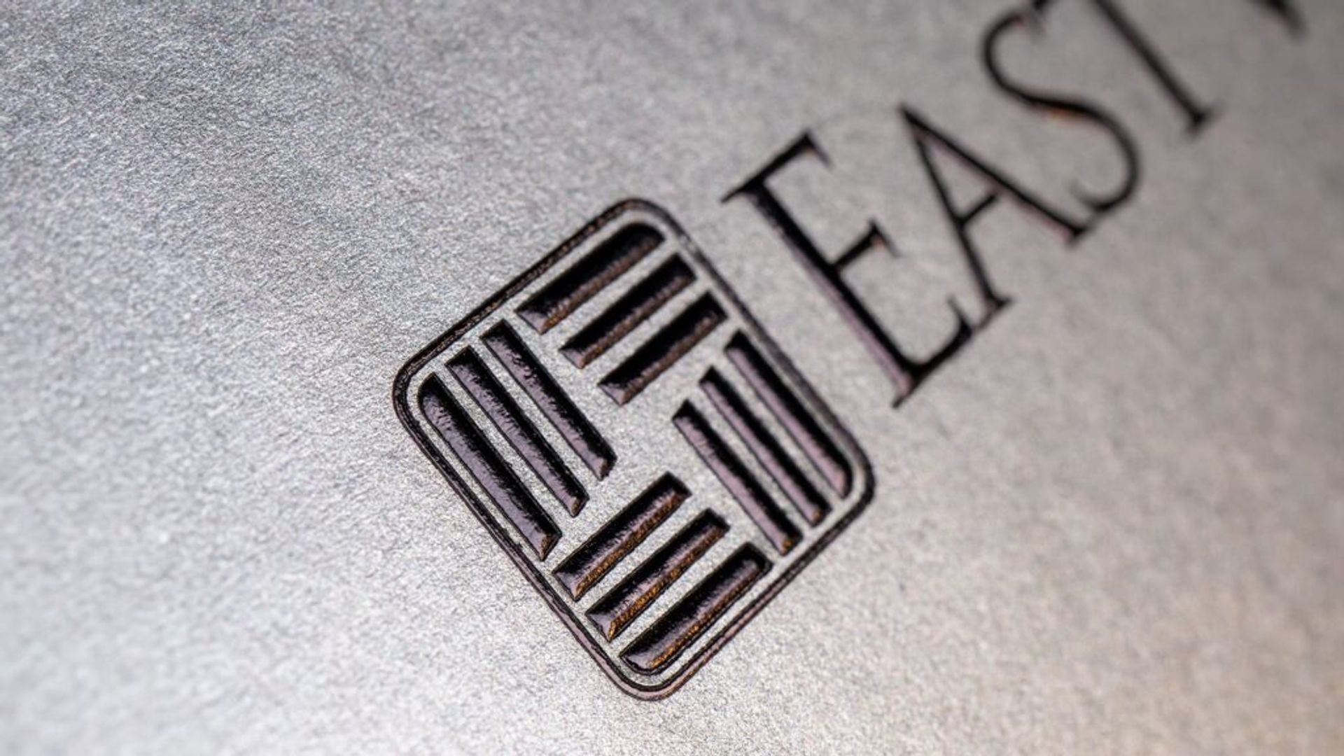 Extreme close-up of the East West Bank geometric emblem and text embossed with glossy black foil on a textured silver metallic background