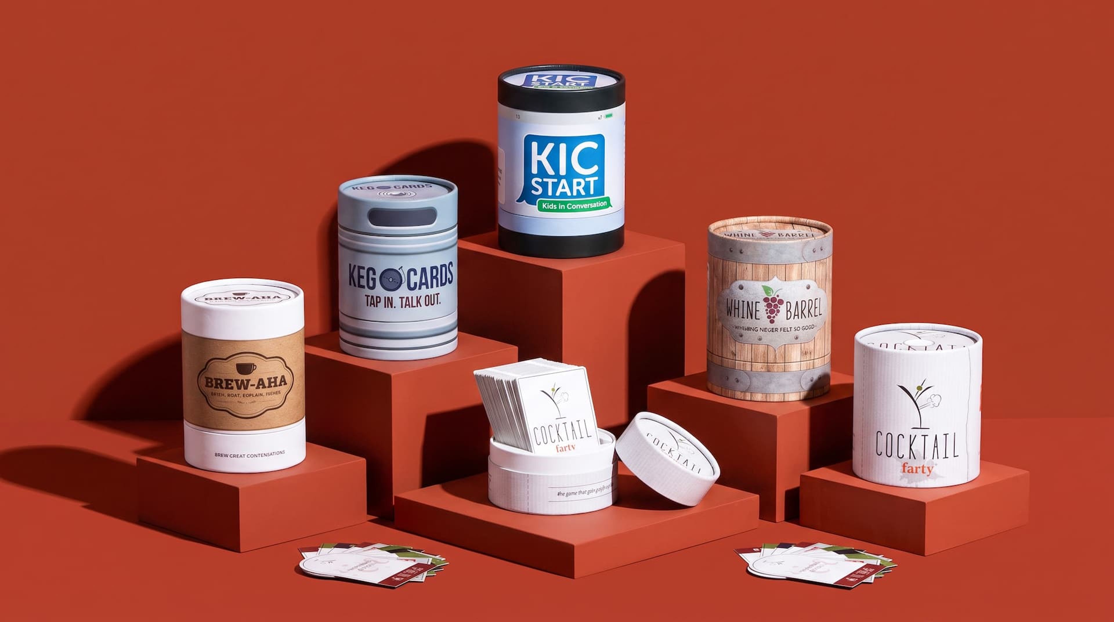 Five cylindrical game containers on terracotta risers. One is open, revealing a stack of cards for "Cocktail Farty."