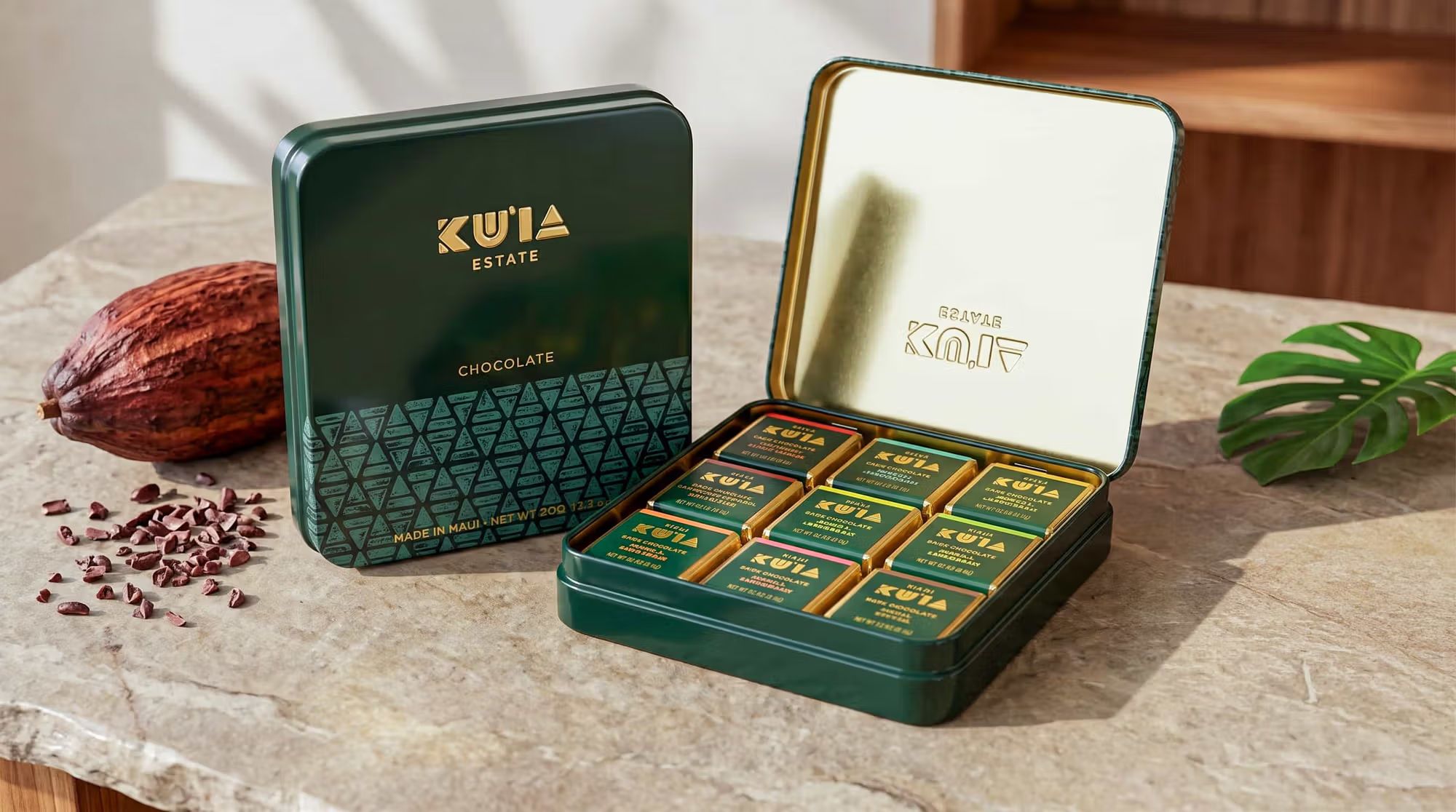 Open and closed green Maui Ku'ia Estate Chocolate tins displaying wrapped square chocolates, a cacao pod, and scattered nibs.