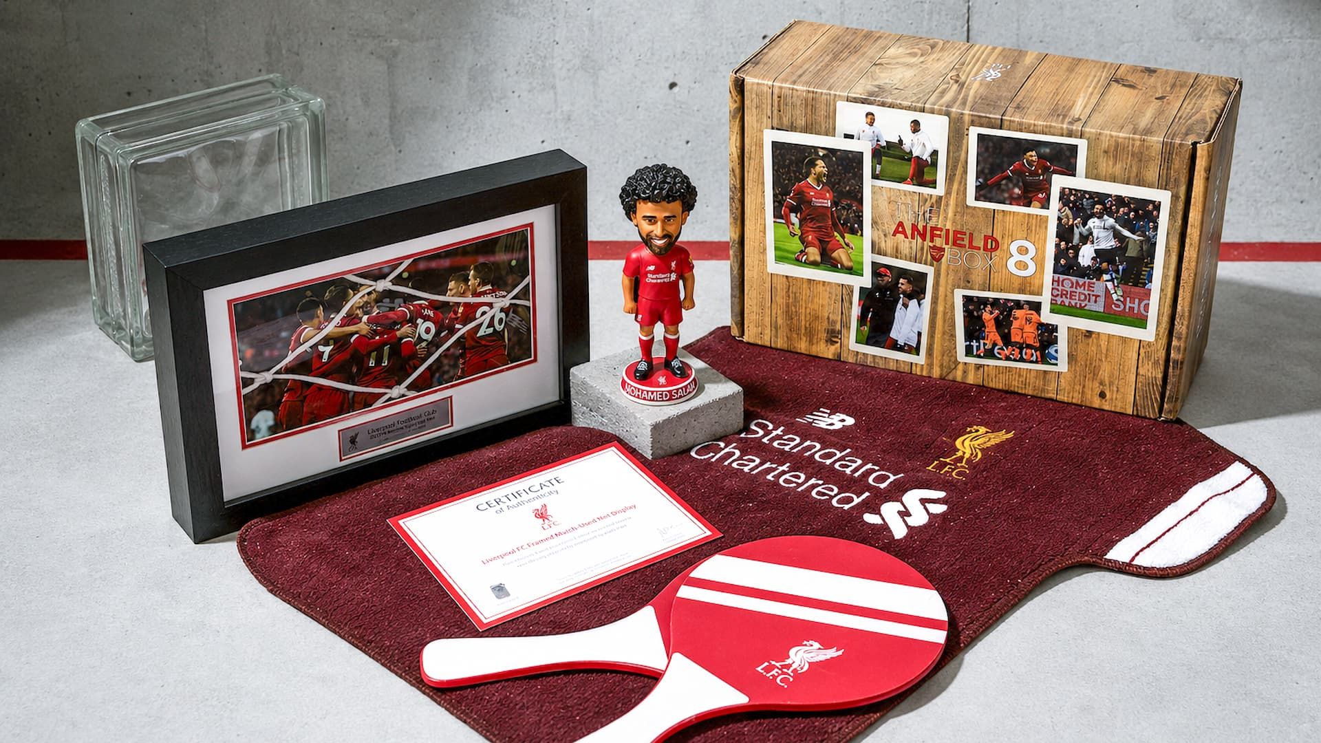 Liverpool FC merchandise displayed outside its wood-grain Anfield Box, including a framed photo, towel, and Mo Salah figure.