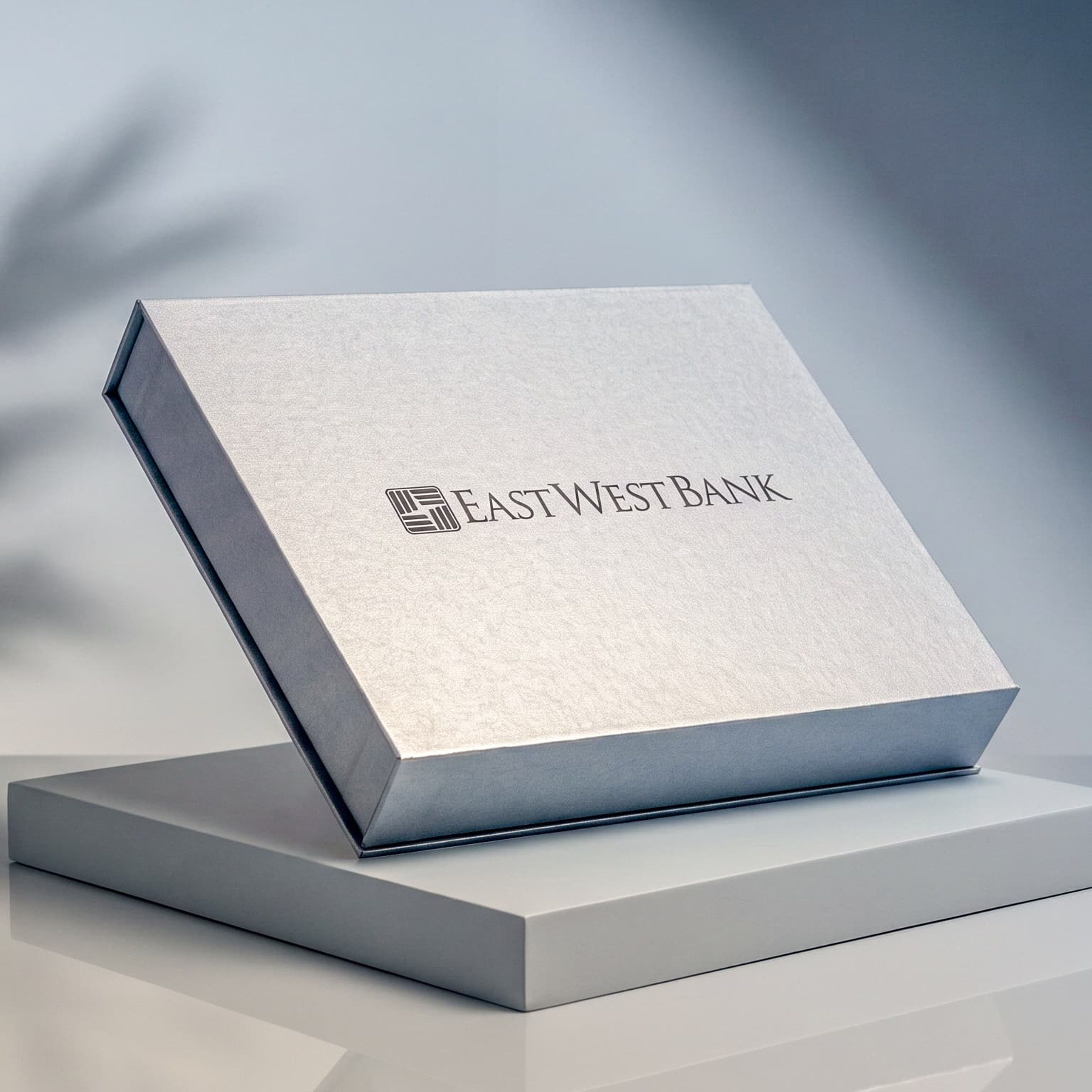 Closed silver East West Bank presentation box propped on a grey pedestal against a wall with leafy plant shadows.