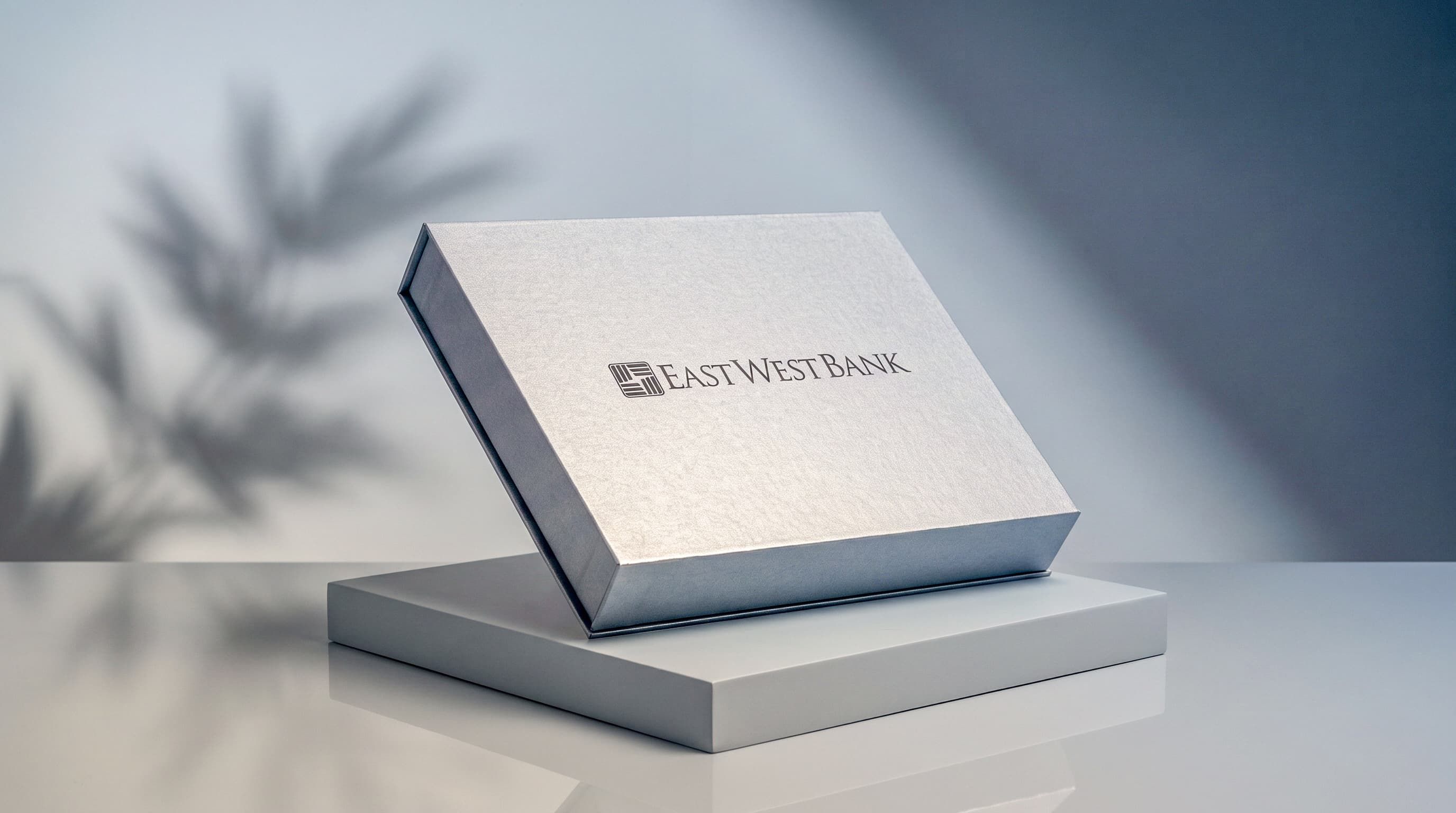 Closed silver East West Bank presentation box propped on a grey pedestal against a wall with leafy plant shadows.