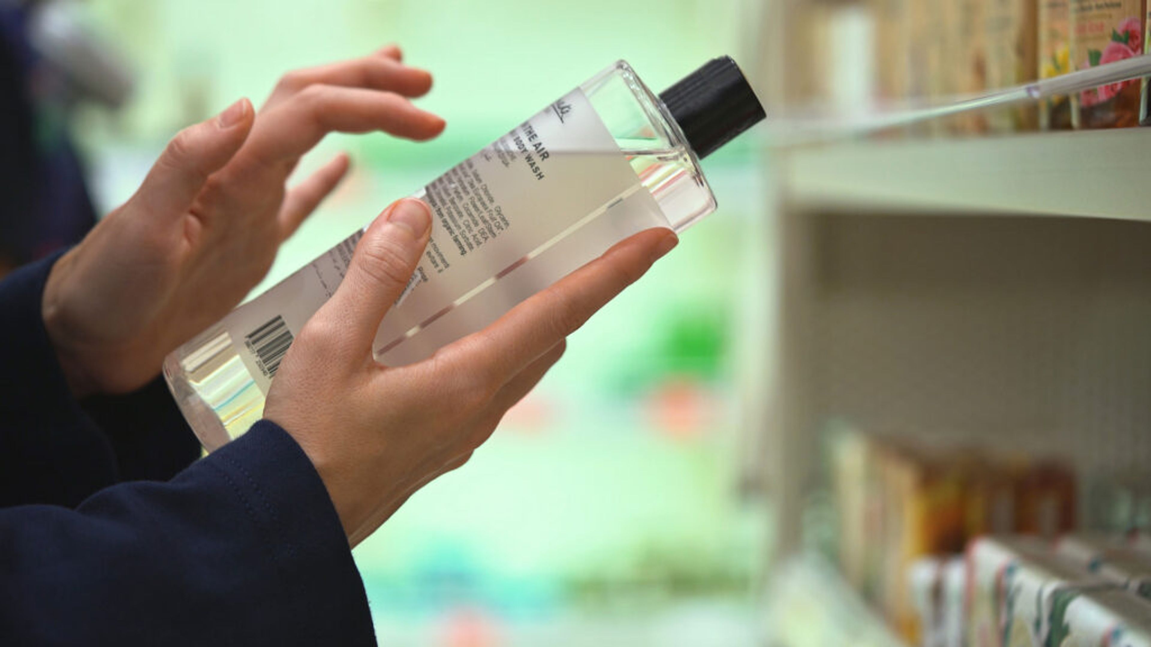 Close-up hands hold transparent bottle of body wash or shampoo. Customer reading label with ingredients, examining composition in cosmetics store, choosing cosmetic product, making purchasing decision