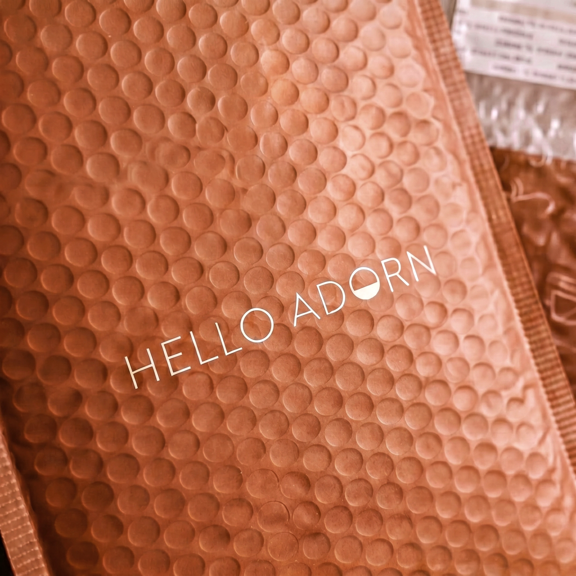 A close-up of a rust-colored bubble mailer with the words "HELLO ADORN" printed in white across the center.