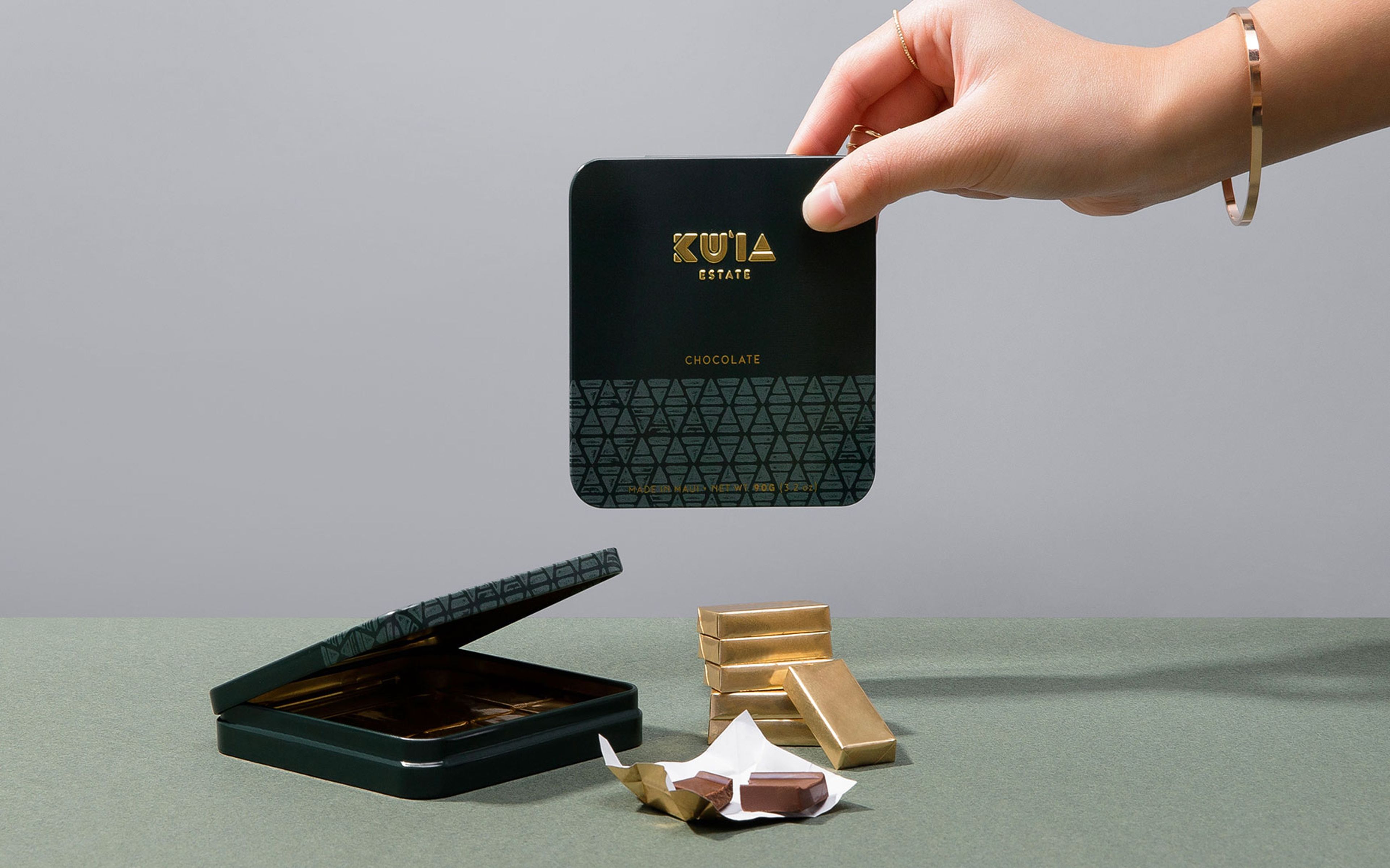 A hand holds a green Ku'ia Estate Chocolate tin over an open tin, gold-foil chocolates, and a loose piece on a green table.